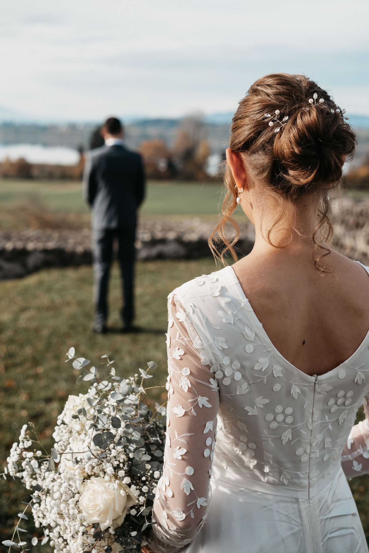 A bride in a delicate lace wedding dress with sheer sleeves holds a white floral bouquet in an outdoor setting.