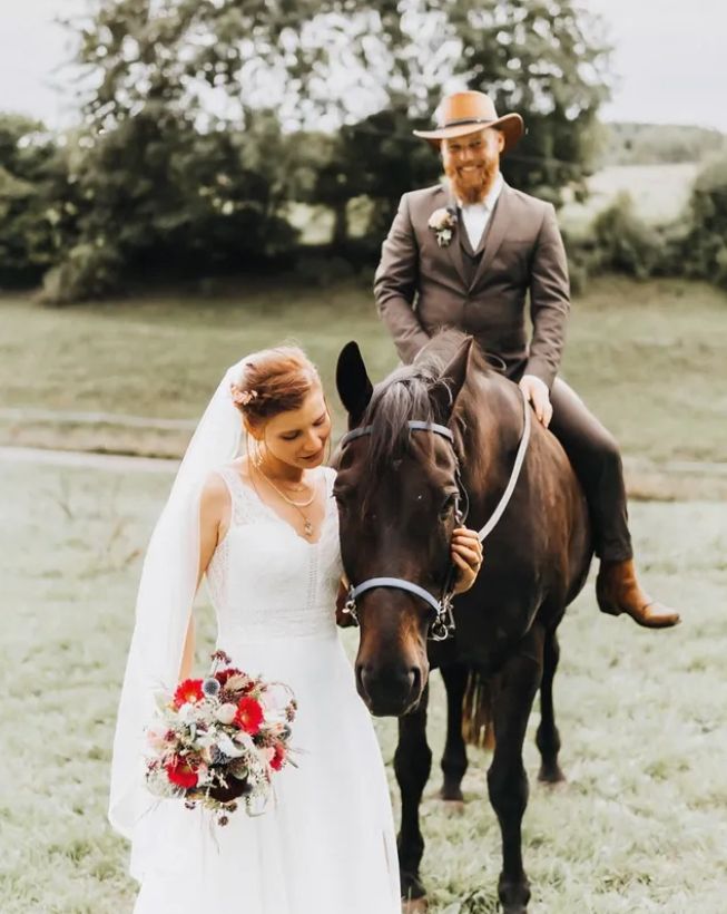 A couple poses with their dark brown horse in a serene outdoor setting during their wedding photoshoot.