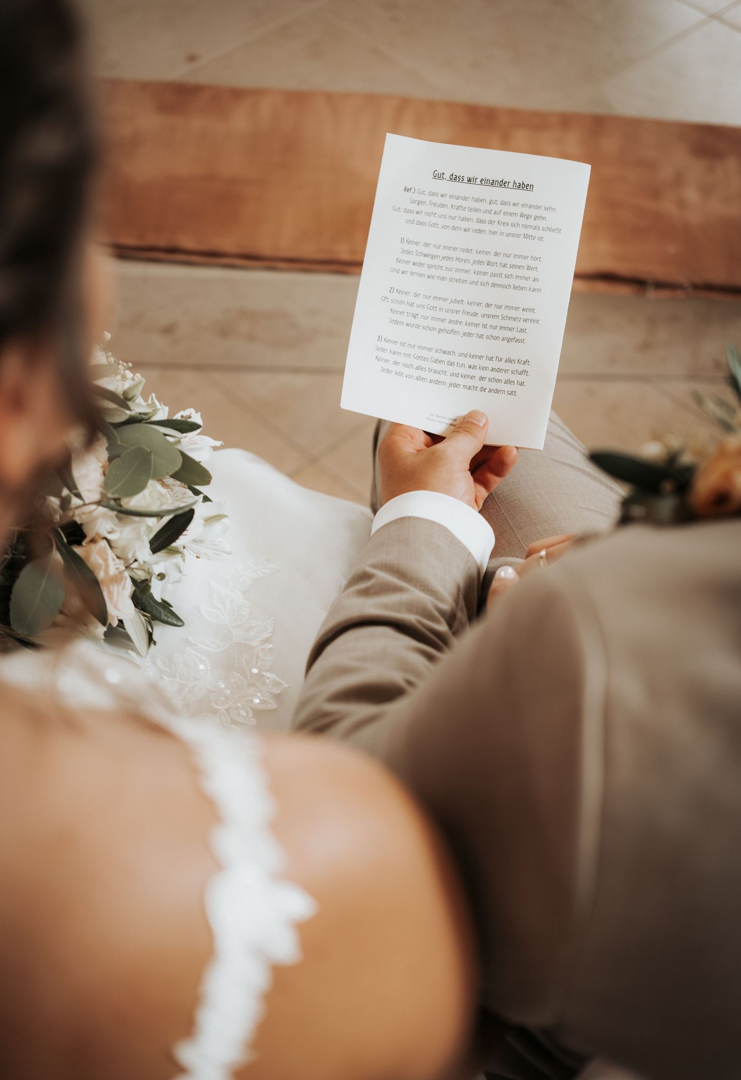 A person holds and reads a white paper note with text while white flowers rest nearby on a beige surface.