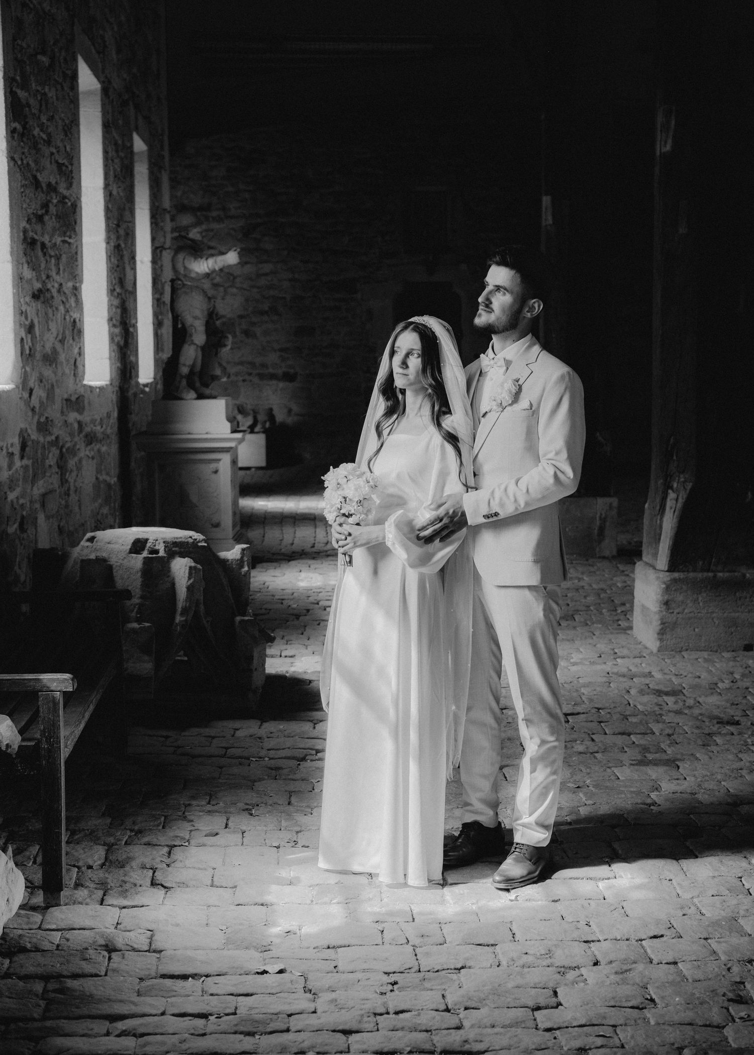 Black and white photo of a bride and groom standing together in a rustic stone chapel, bride holding a bouquet.
