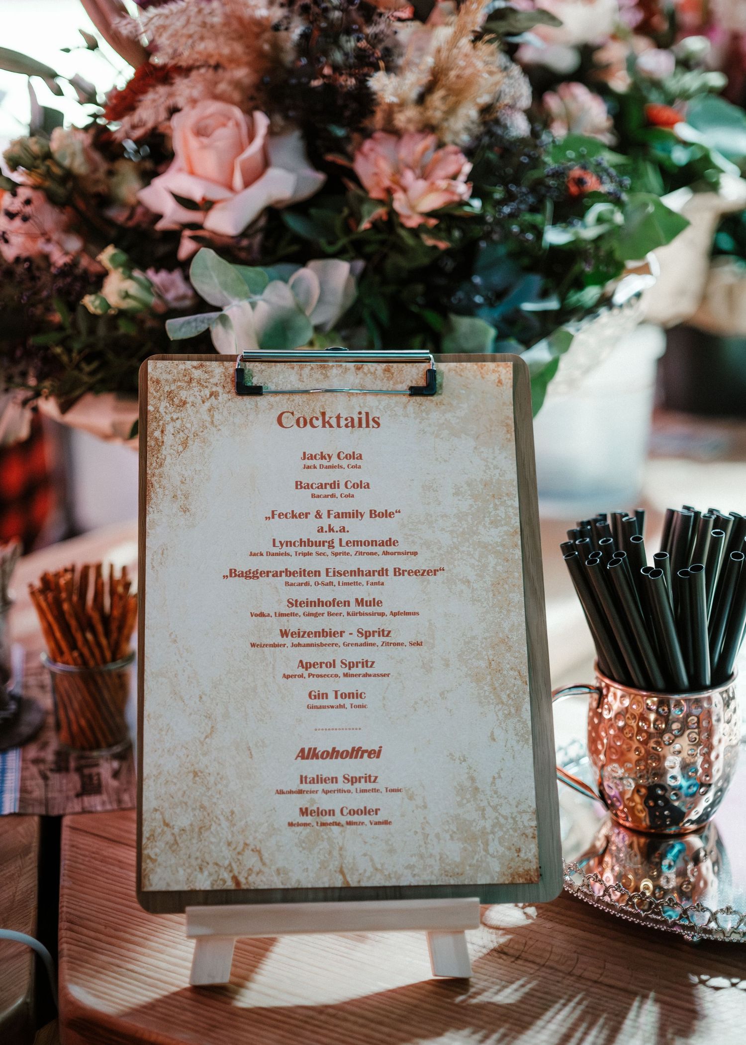 A vintage-style menu card displayed at an event with floral arrangements and black cocktail straws in glass holders.