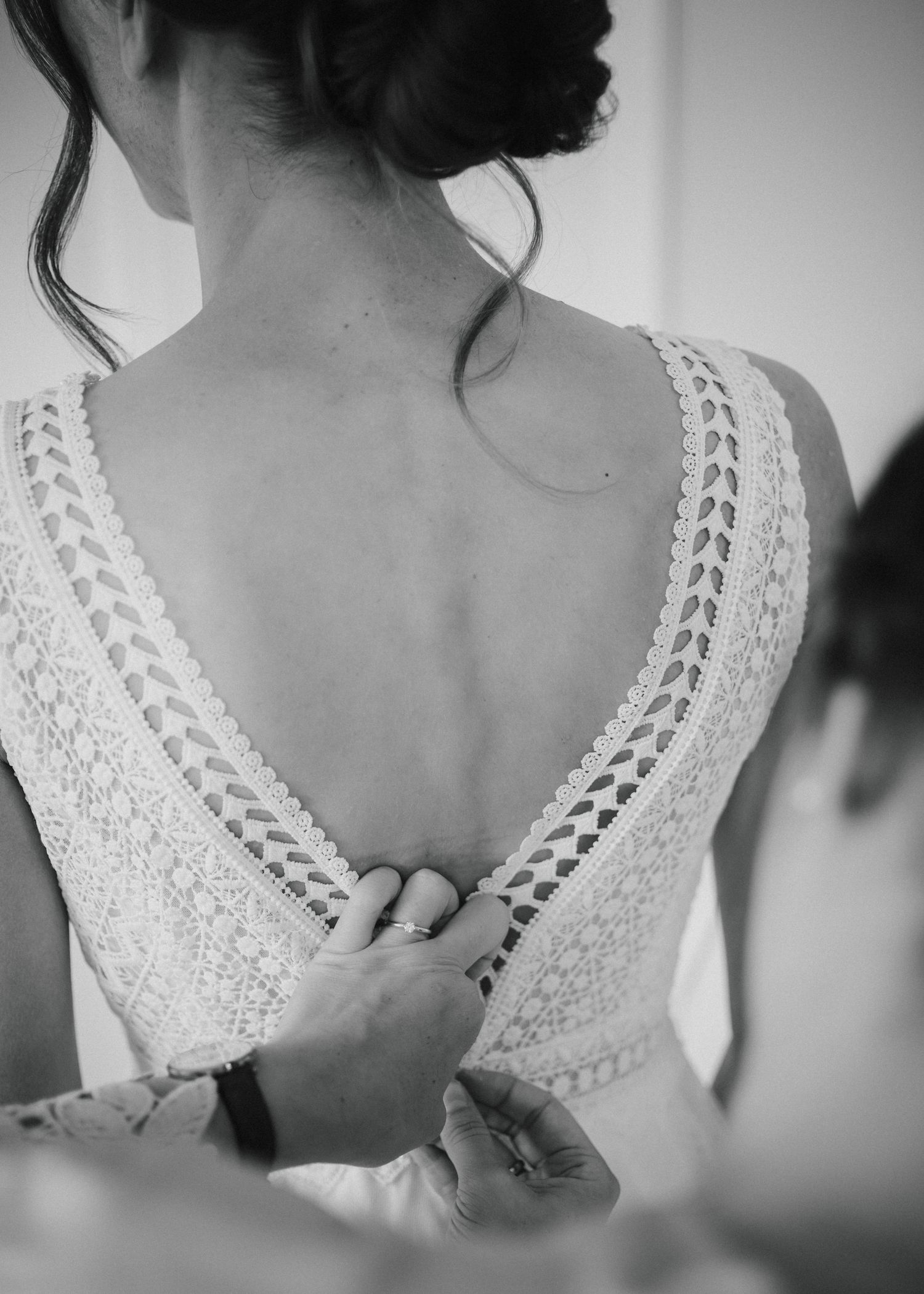 Black and white photo of a bride in a lace backless wedding dress being buttoned up by a helper.