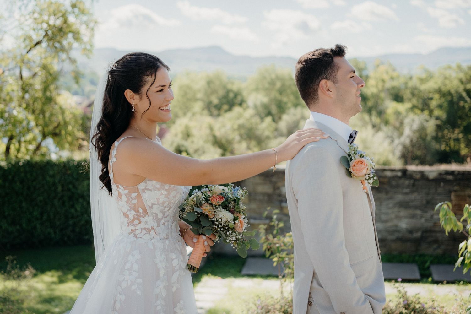 Bride in lace gown surprises groom in tan suit during outdoor first look at wedding ceremony.