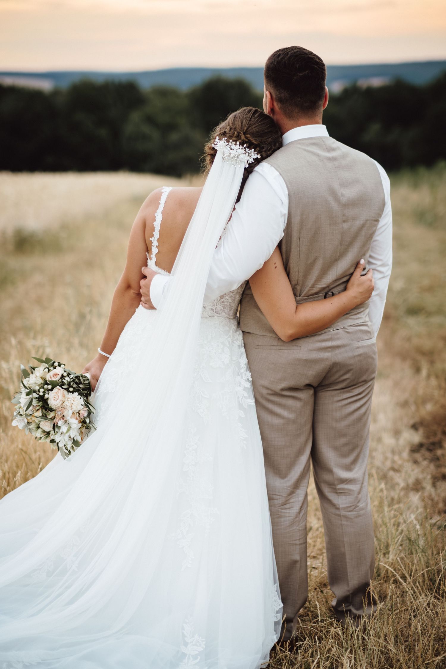 A couple in wedding attire stands together in a golden field at sunset, with the bride holding a white floral bouquet.