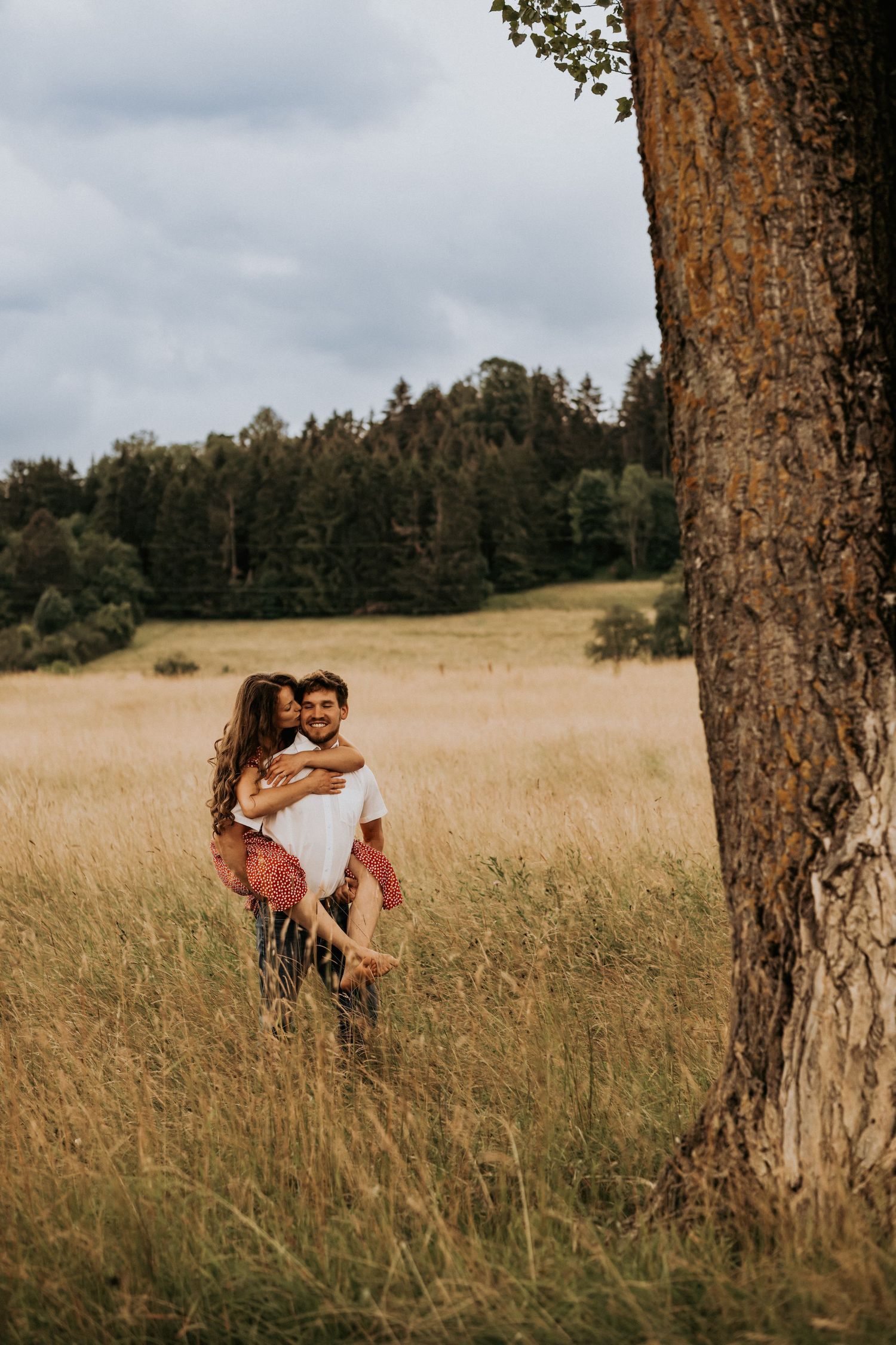 A couple embraces in a golden field of tall grass with trees and cloudy skies in the background.