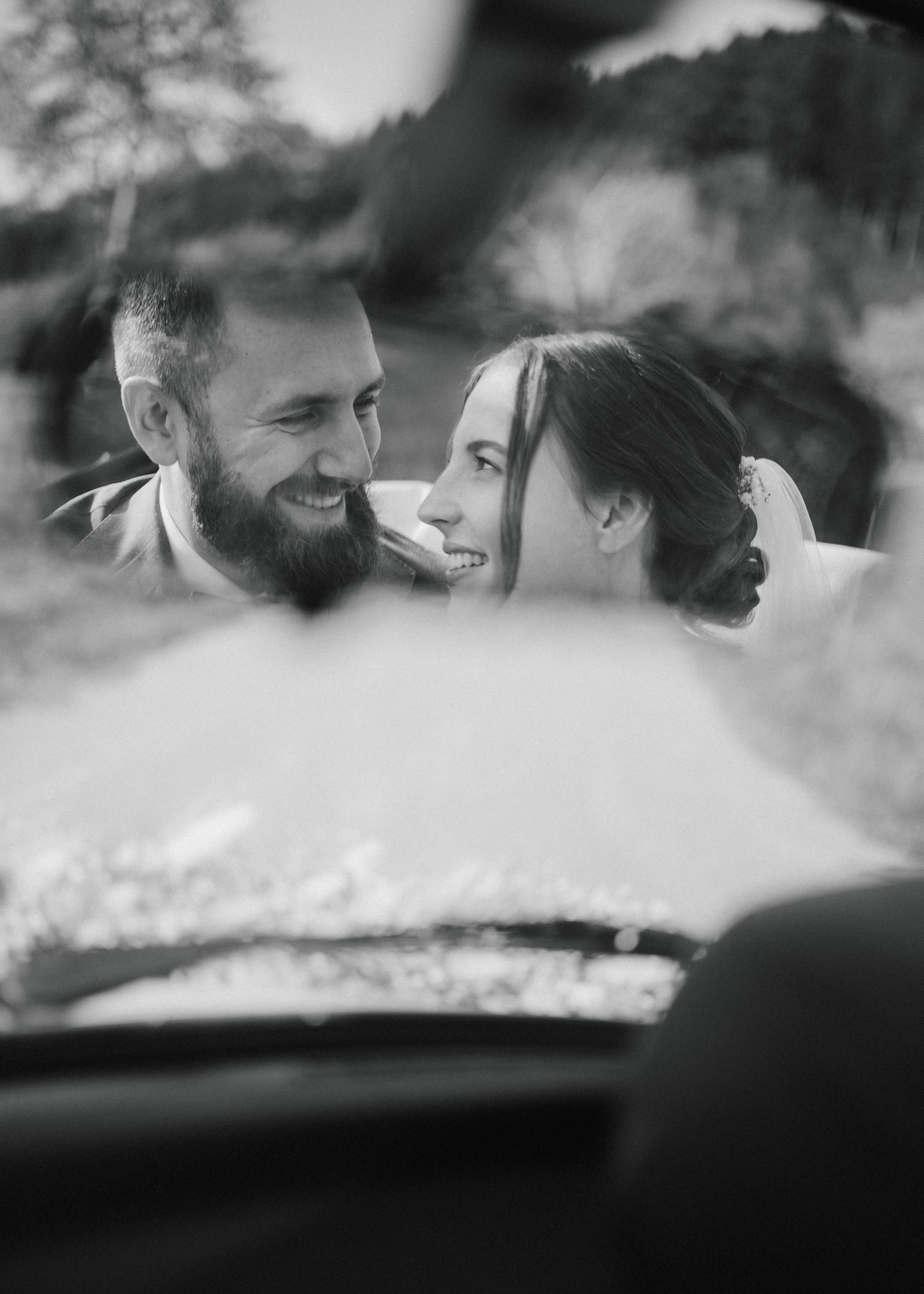 Black and white photo of a smiling bearded groom and bride gazing at each other, shot through a car window.