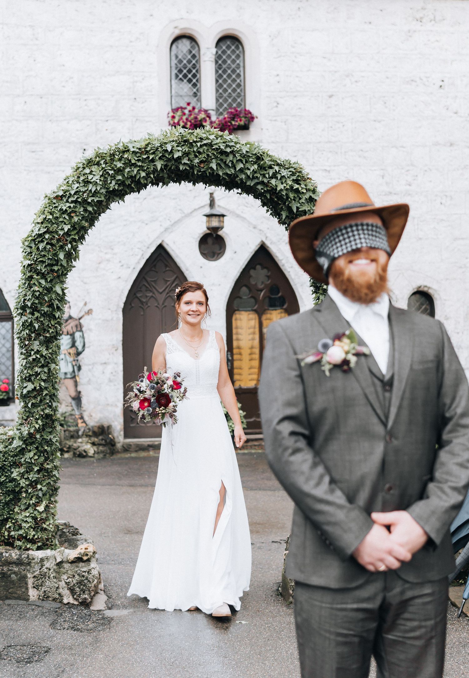 A wedding couple poses under ivy-covered archways against a white stone church wall.