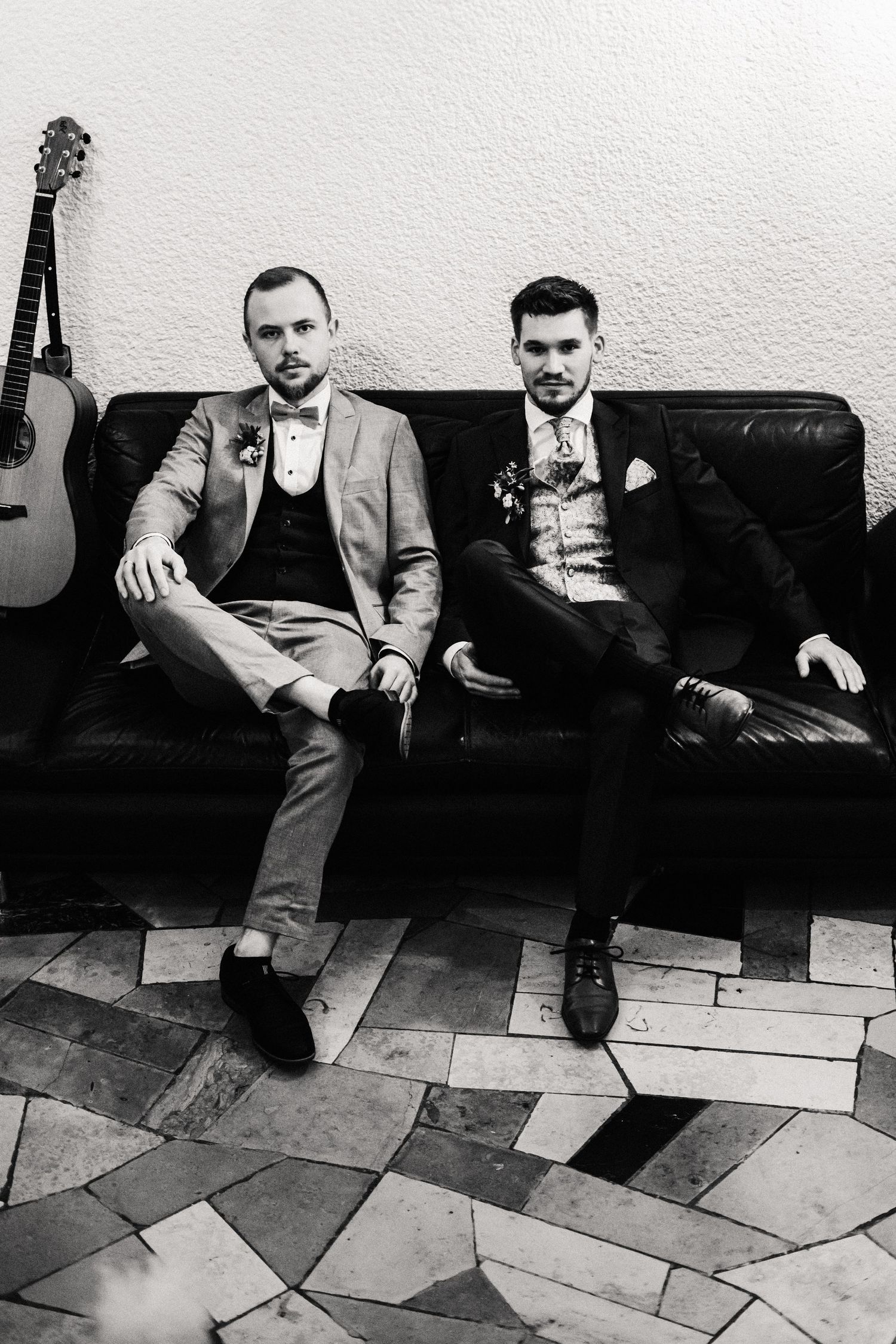 Black and white photo of two men in formal suits sitting on a leather couch with a guitar mounted on the wall.