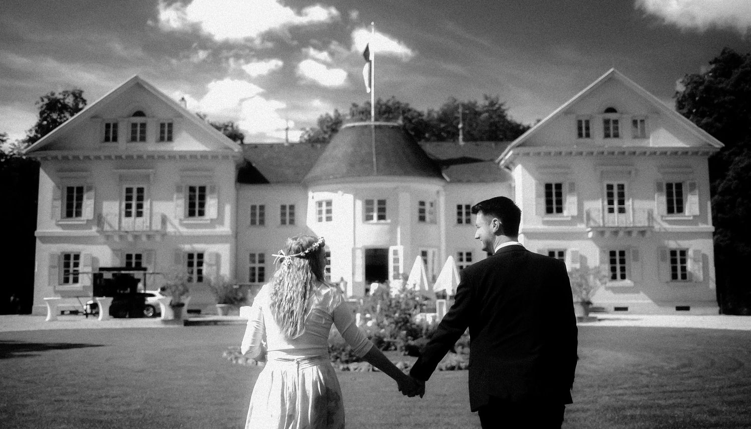 A black and white photograph of a couple holding hands in front of a grand white mansion on a cloudy day.