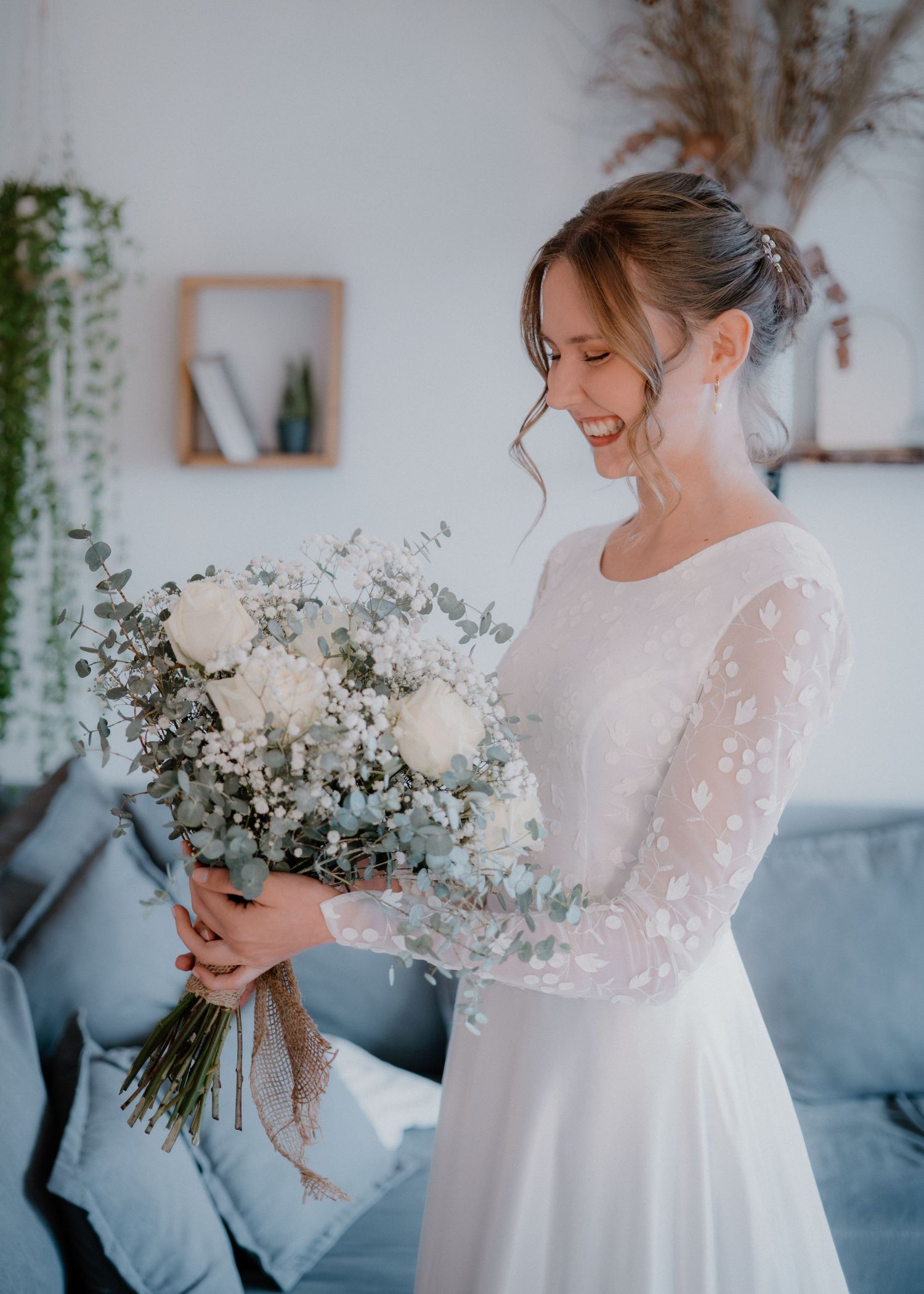 A bride in a white long-sleeved dress holds a delicate bouquet of white and eucalyptus flowers in a bright interior space.