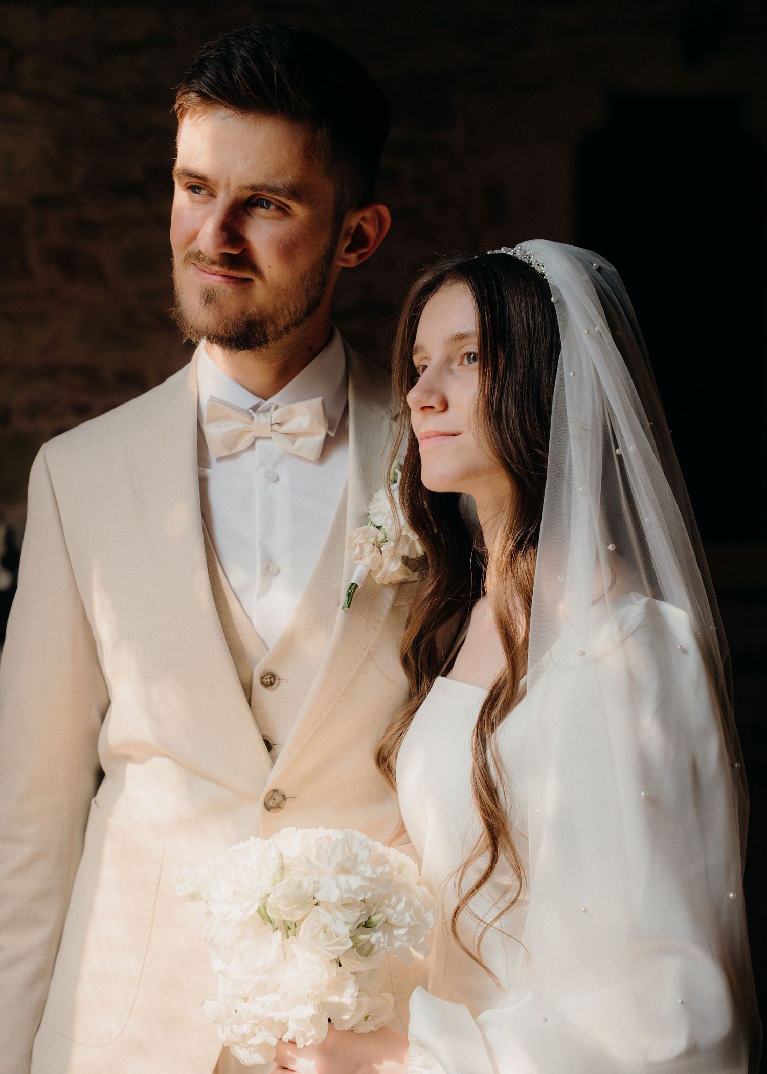 Elegant bride and groom in white wedding attire pose together, bride holding white bouquet with veil, groom in cream suit.