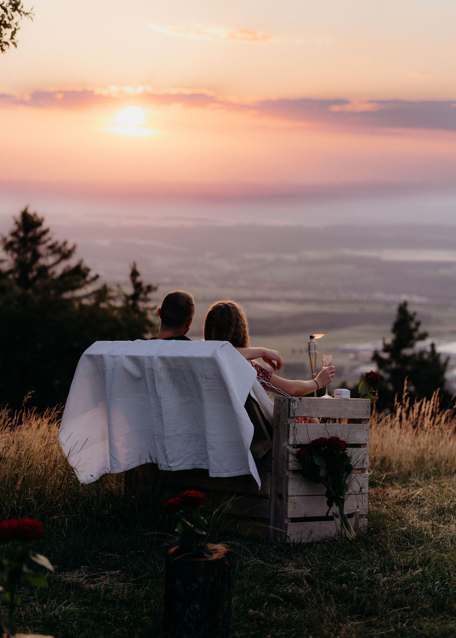 A couple enjoys a romantic sunset picnic on a hilltop overlooking a misty valley with evergreen trees in the foreground.