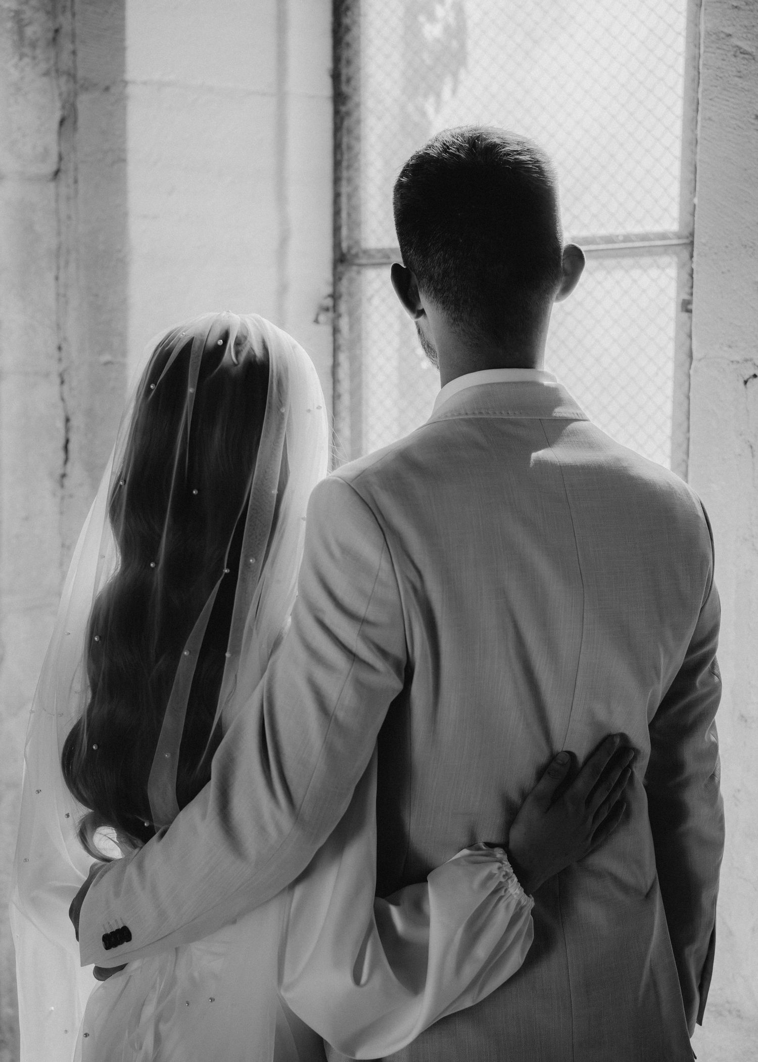 Black and white photo of a bride and groom standing together, viewed from behind, near a window.