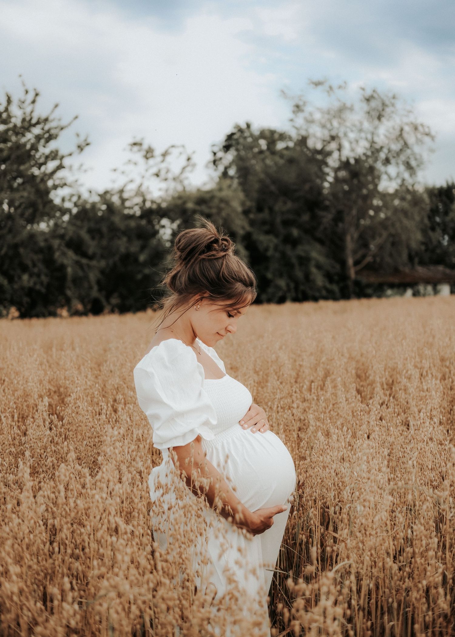 A woman in a white dress stands in a golden wheat field during sunset creating a dreamy maternity photoshoot.