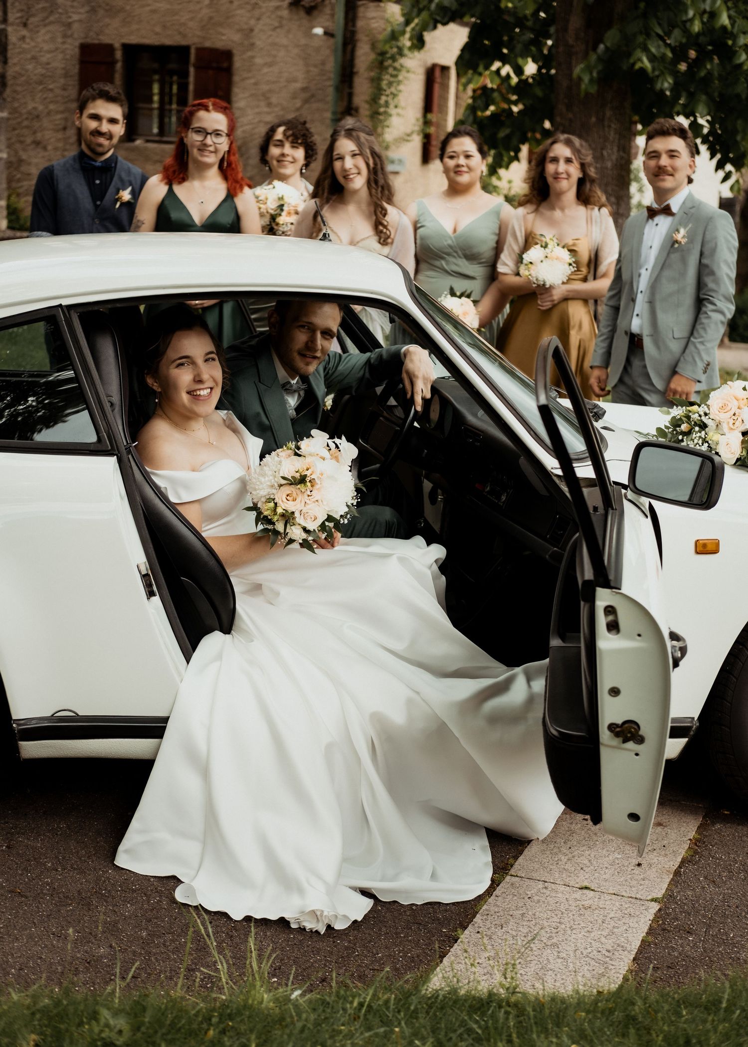 A bride in a white dress entering a classic car while wedding guests look on.