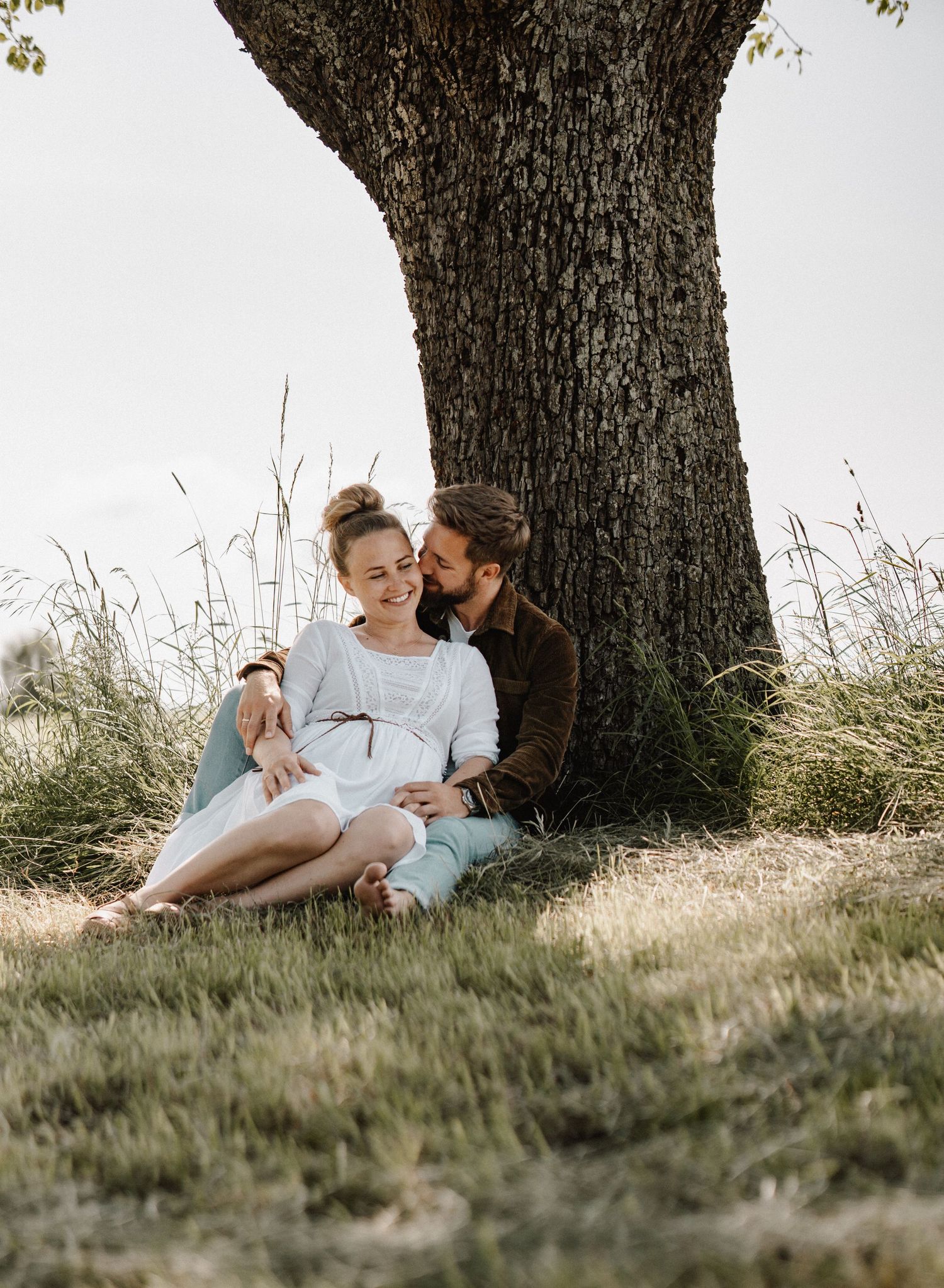 A couple relaxes together under a large tree on a grassy hillside during a warm summer day.