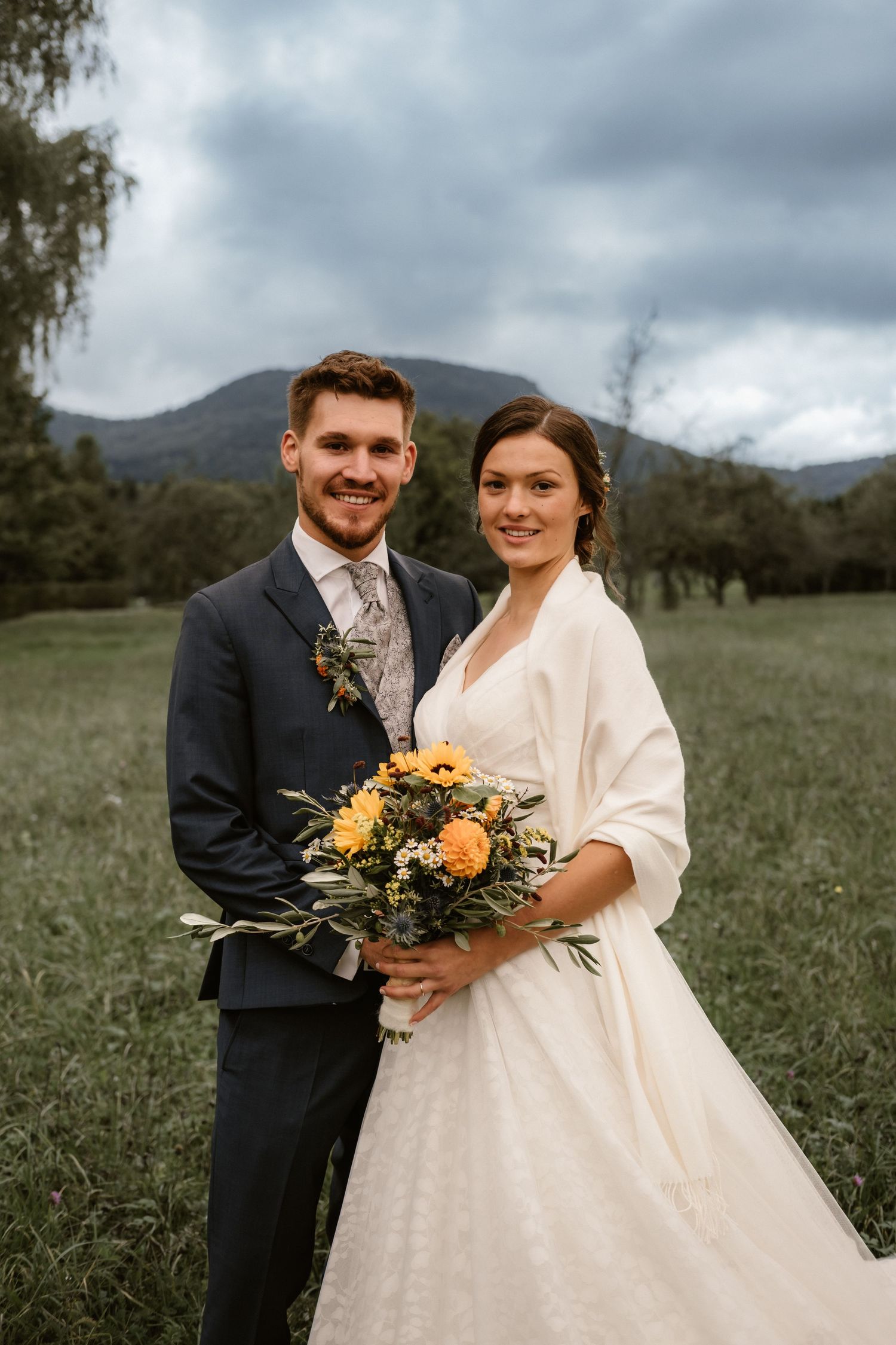 A couple poses together in an outdoor field with mountains in the background during their wedding ceremony.