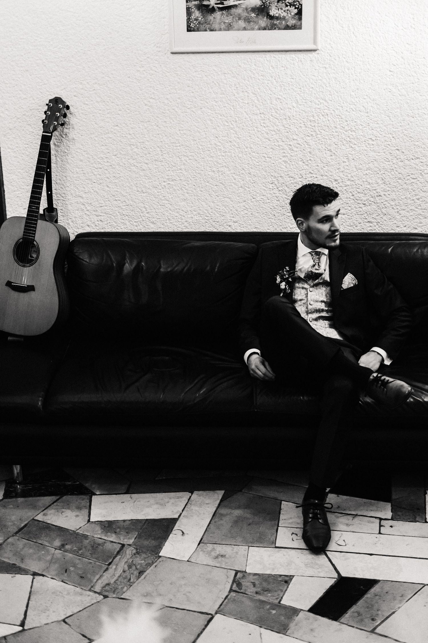 Black and white photo of someone in formal attire sitting on a leather couch with an acoustic guitar mounted on the wall.