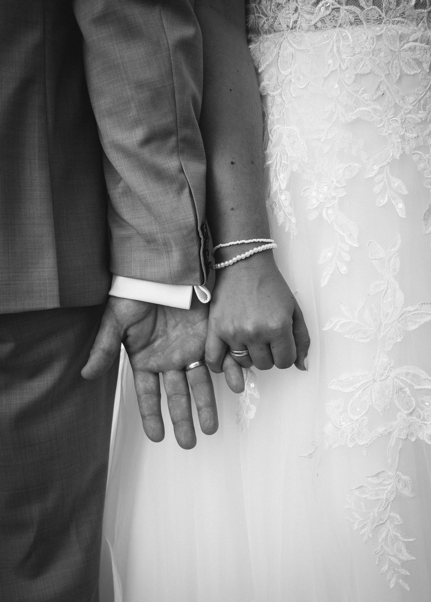 Close-up black and white photo of newlyweds' hands showing their wedding rings against a white lace dress.
