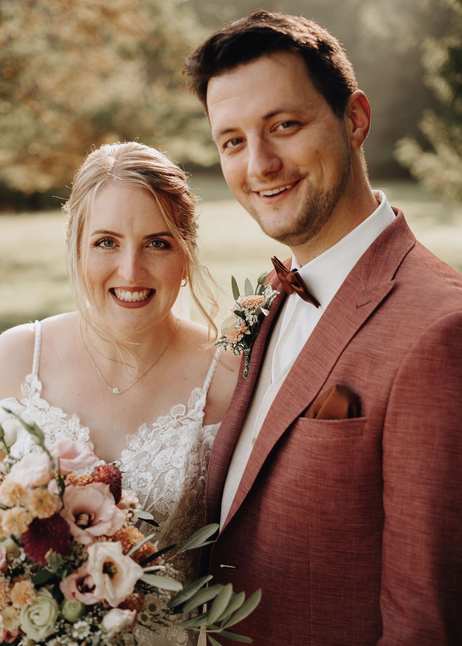 A couple in wedding attire poses together outdoors, with the bride in white lace and groom in burgundy suit.
