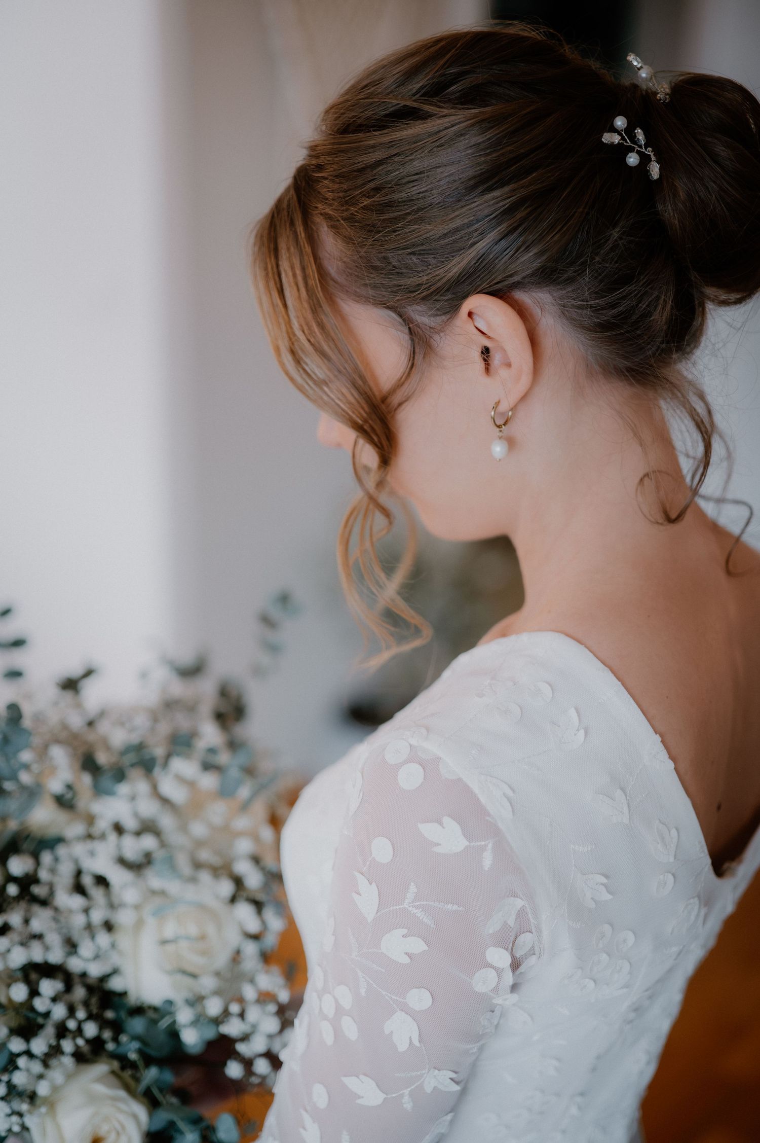 Bride in delicate lace wedding dress shown in artistic profile view with soft floral decorations in background.