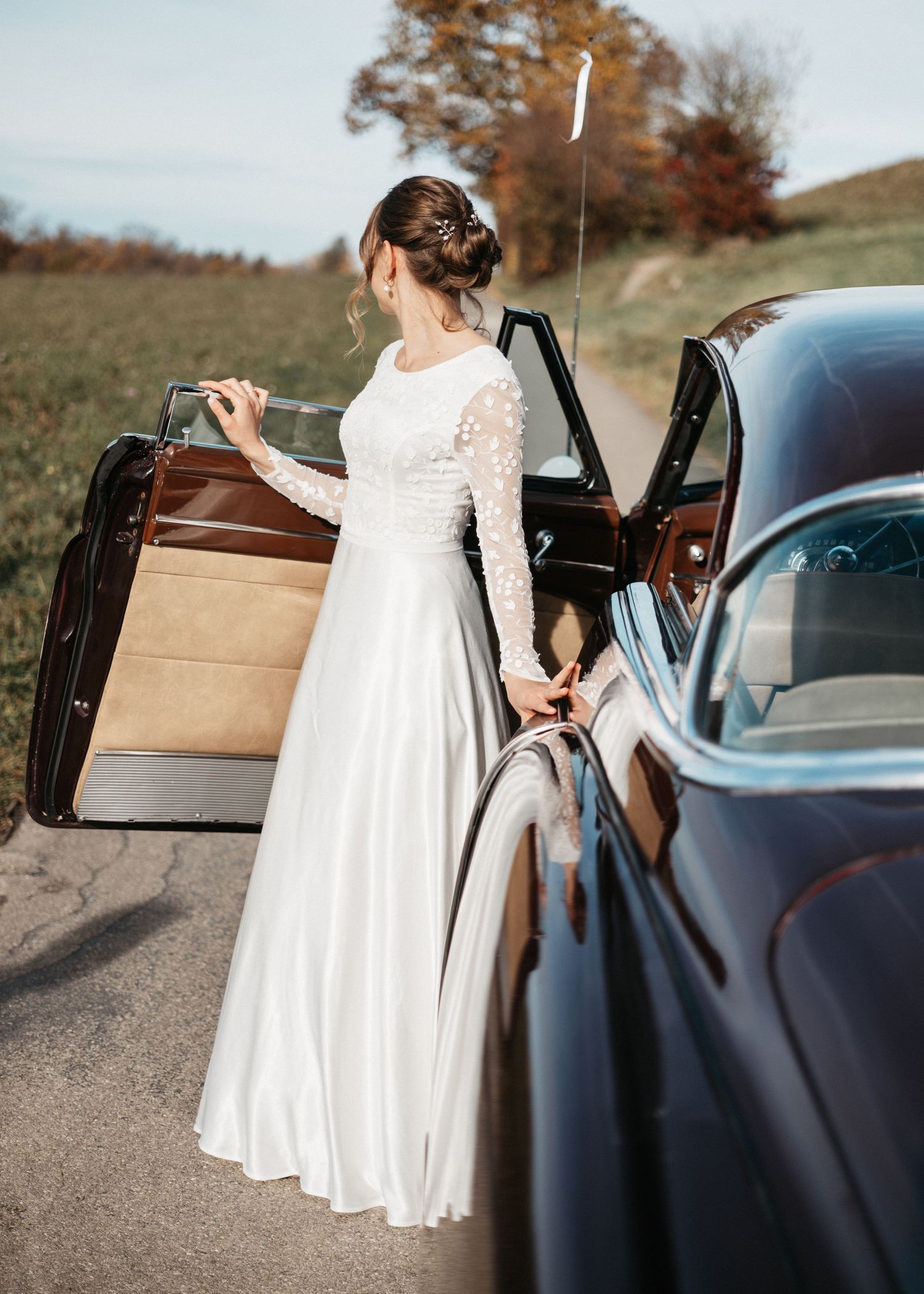 A person in a white dress stands beside a parked car in an outdoor setting during autumn.
