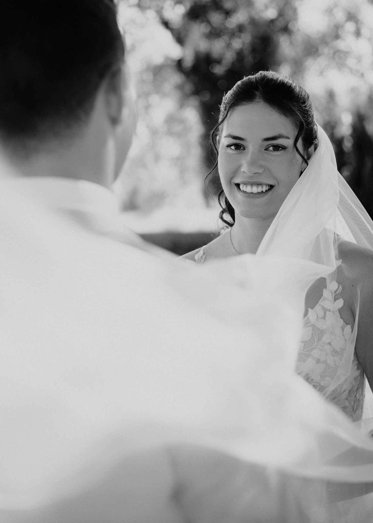 Black and white wedding photo of a smiling bride in veil gazing at groom during outdoor ceremony.