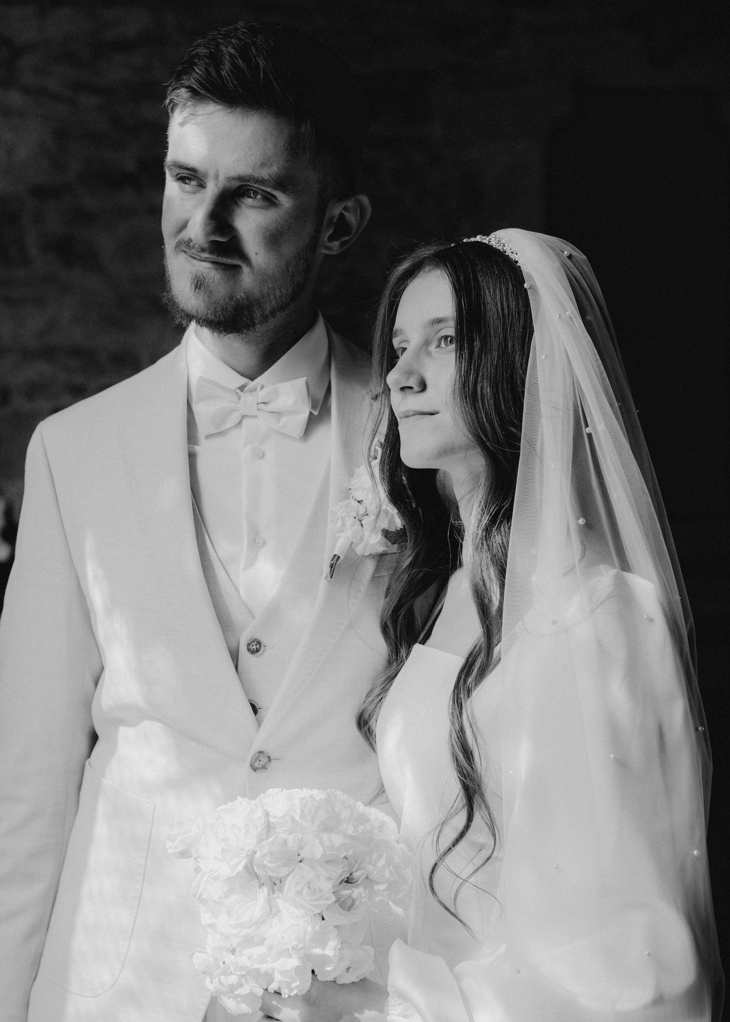 Black and white wedding portrait of bride holding bouquet and groom in white suit, gazing lovingly together.