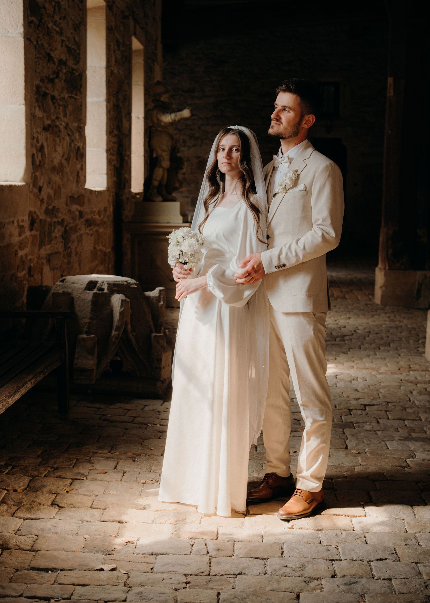 Bride in white gown and groom in cream suit stand together in a rustic stone venue, bathed in warm light.