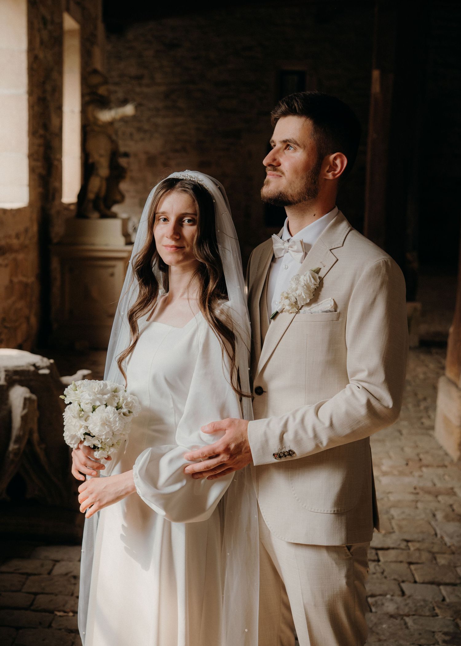 Elegant bride and groom in cream wedding attire pose together in a rustic stone venue holding white flowers.