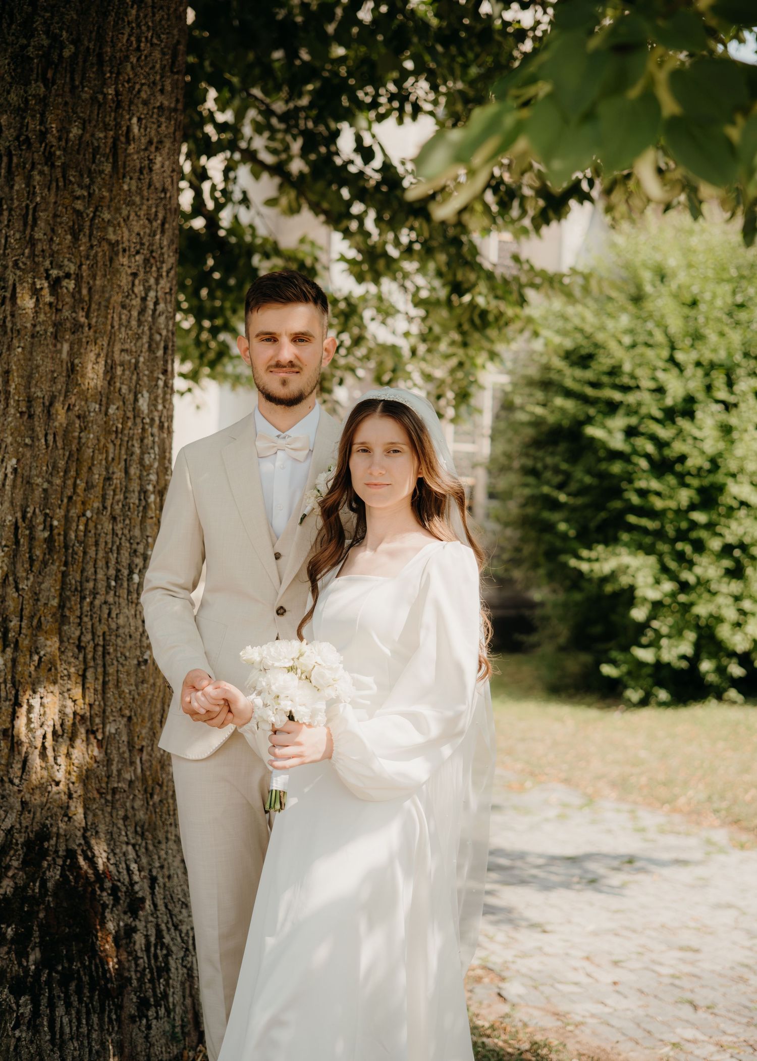 A couple in wedding attire poses by a tree, the bride holding white flowers in a flowing gown, groom in a beige suit.