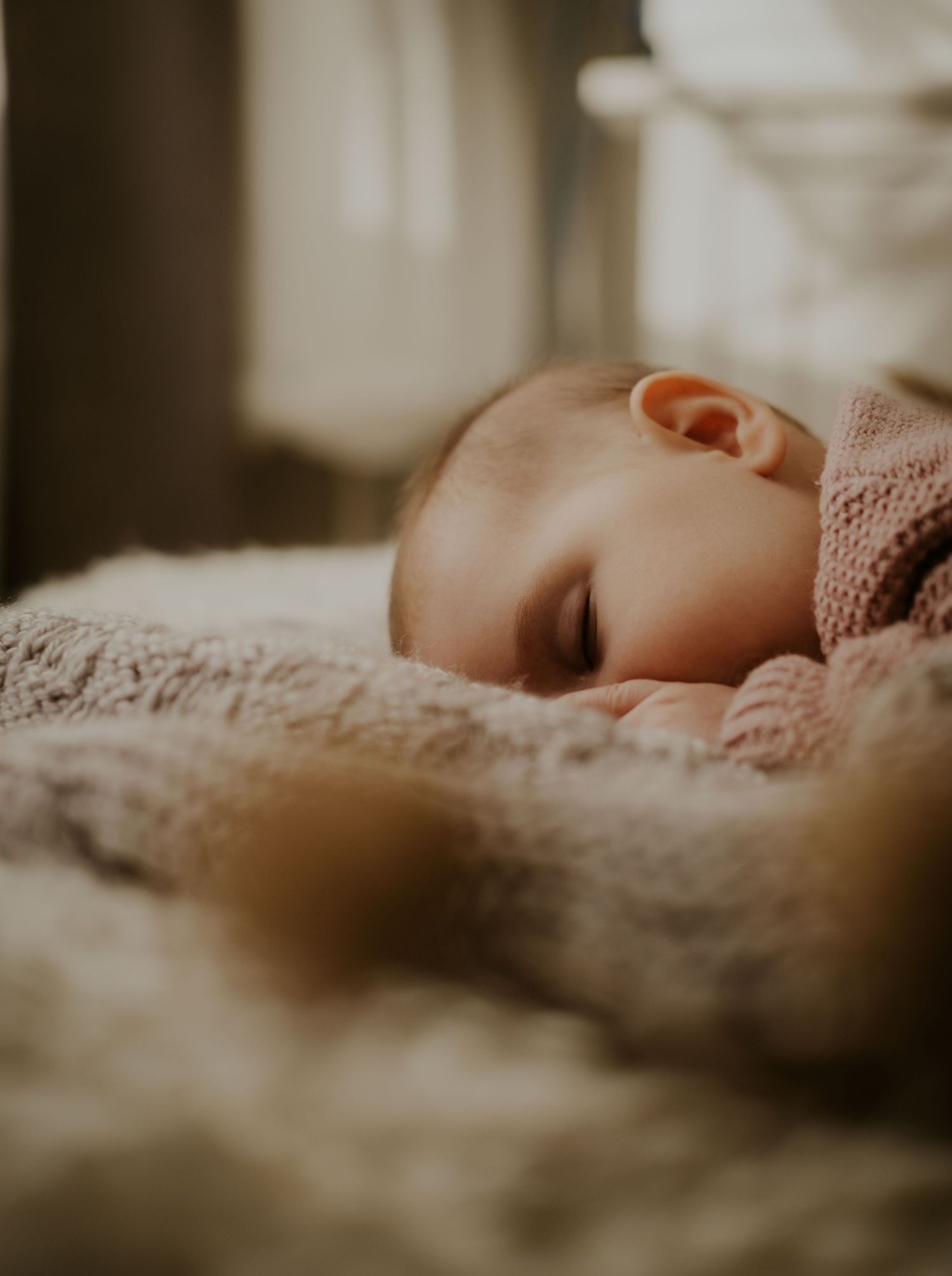 A baby sleeps peacefully on soft beige lace bedding in a dreamy, muted light setting.