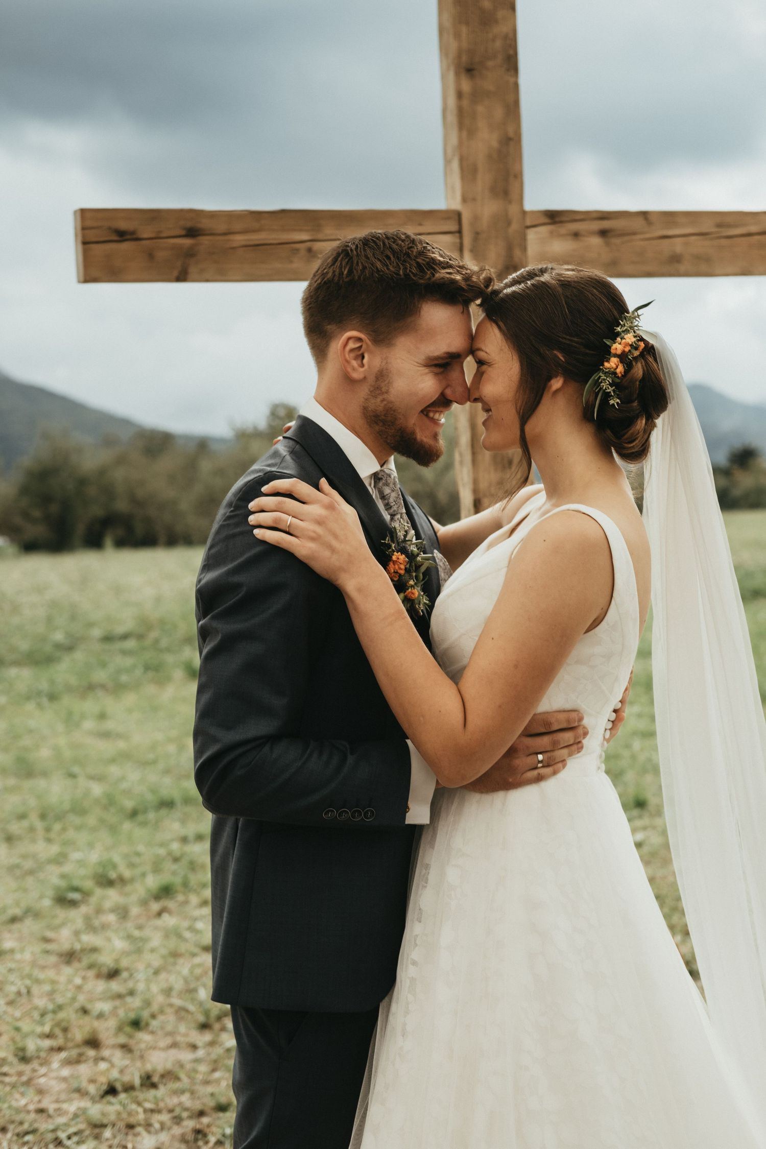 A couple embraces during their outdoor wedding ceremony in front of a wooden cross with mountains in the background.