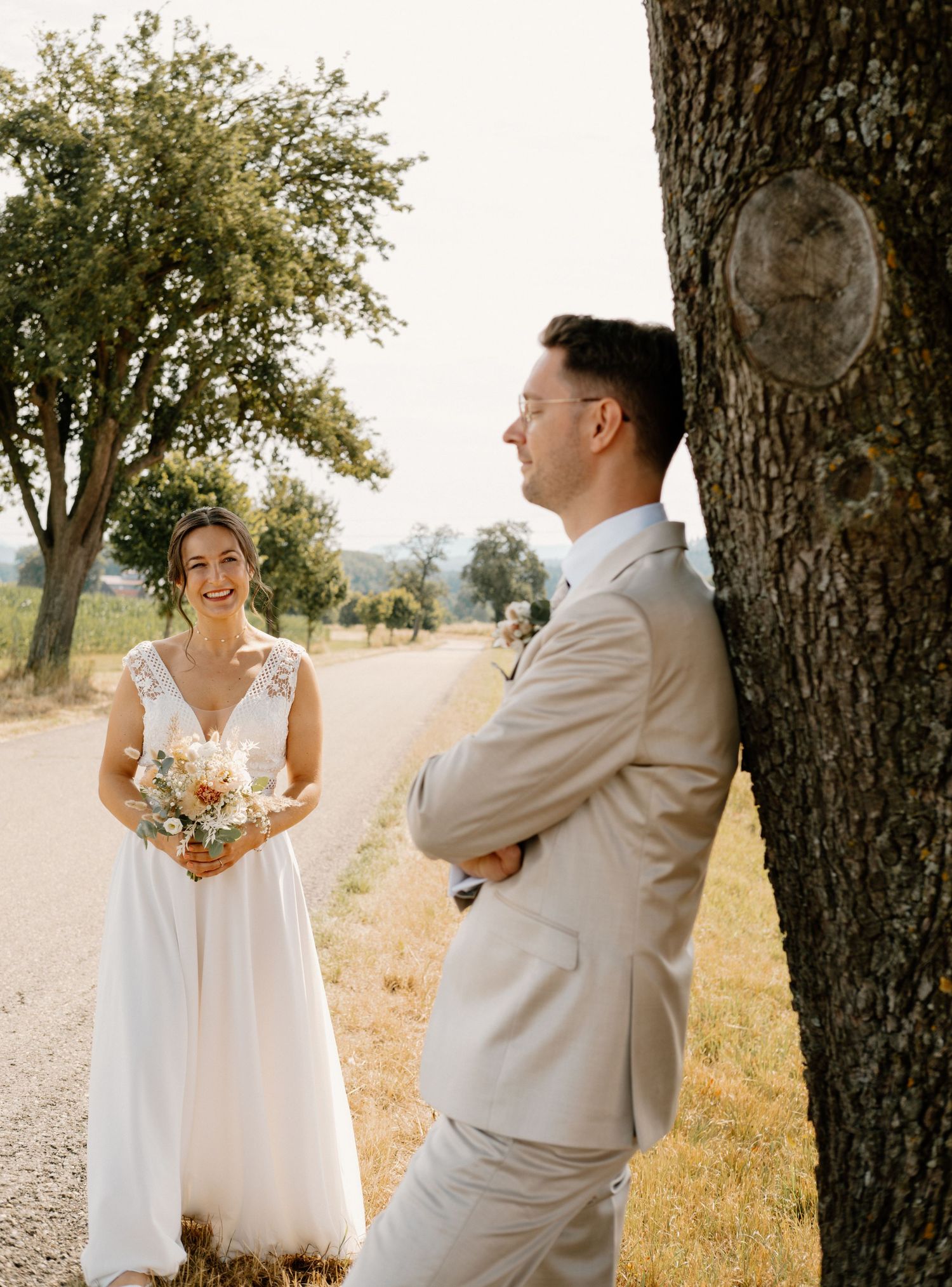 A couple in wedding attire shares a joyful first look moment on a sunny dirt road with trees in the background.