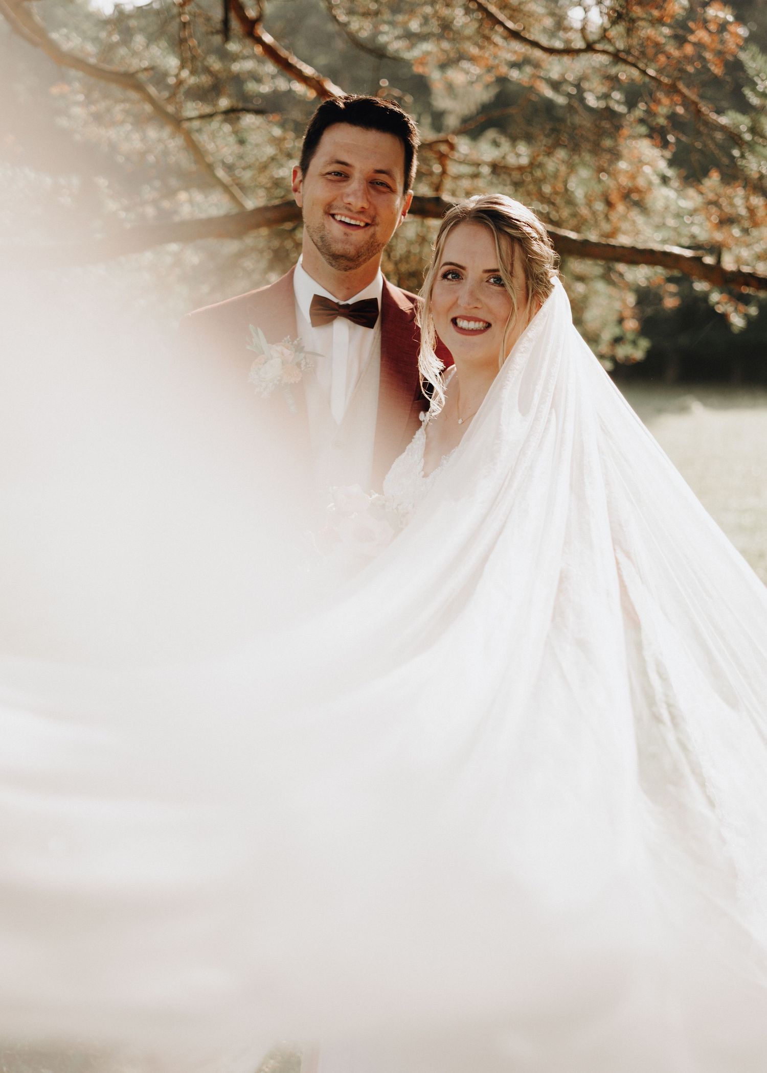 A couple in wedding attire embraces under autumn foliage during their outdoor photoshoot.