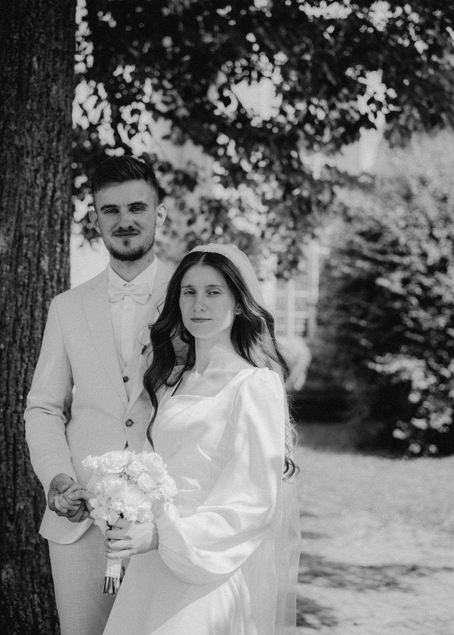 Black and white wedding photo of a bride holding flowers and groom in suit posing near a tree outdoors.