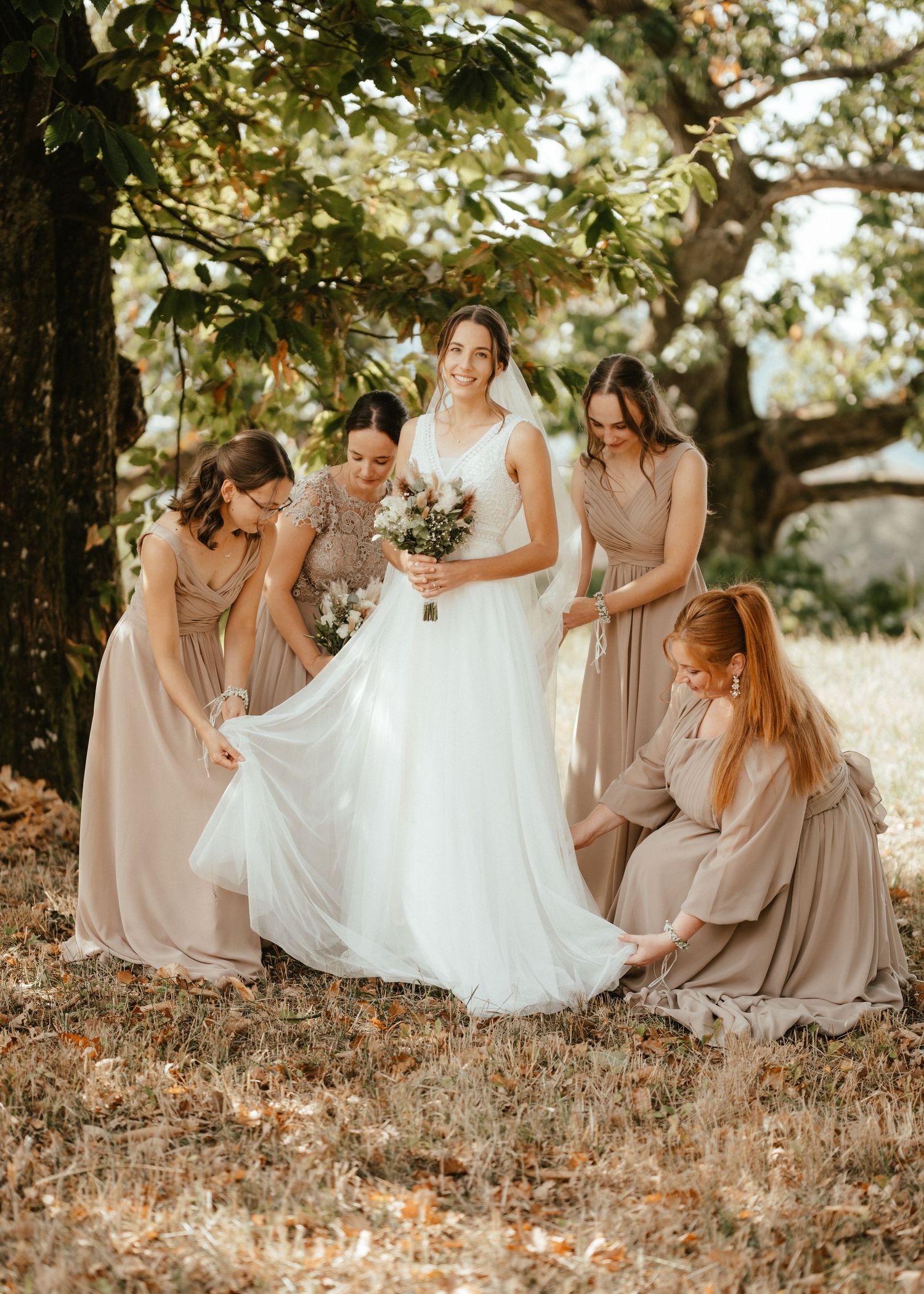 Bride in white gown holds bouquet as four bridesmaids in taupe dresses arrange her train outdoors under a tree.
