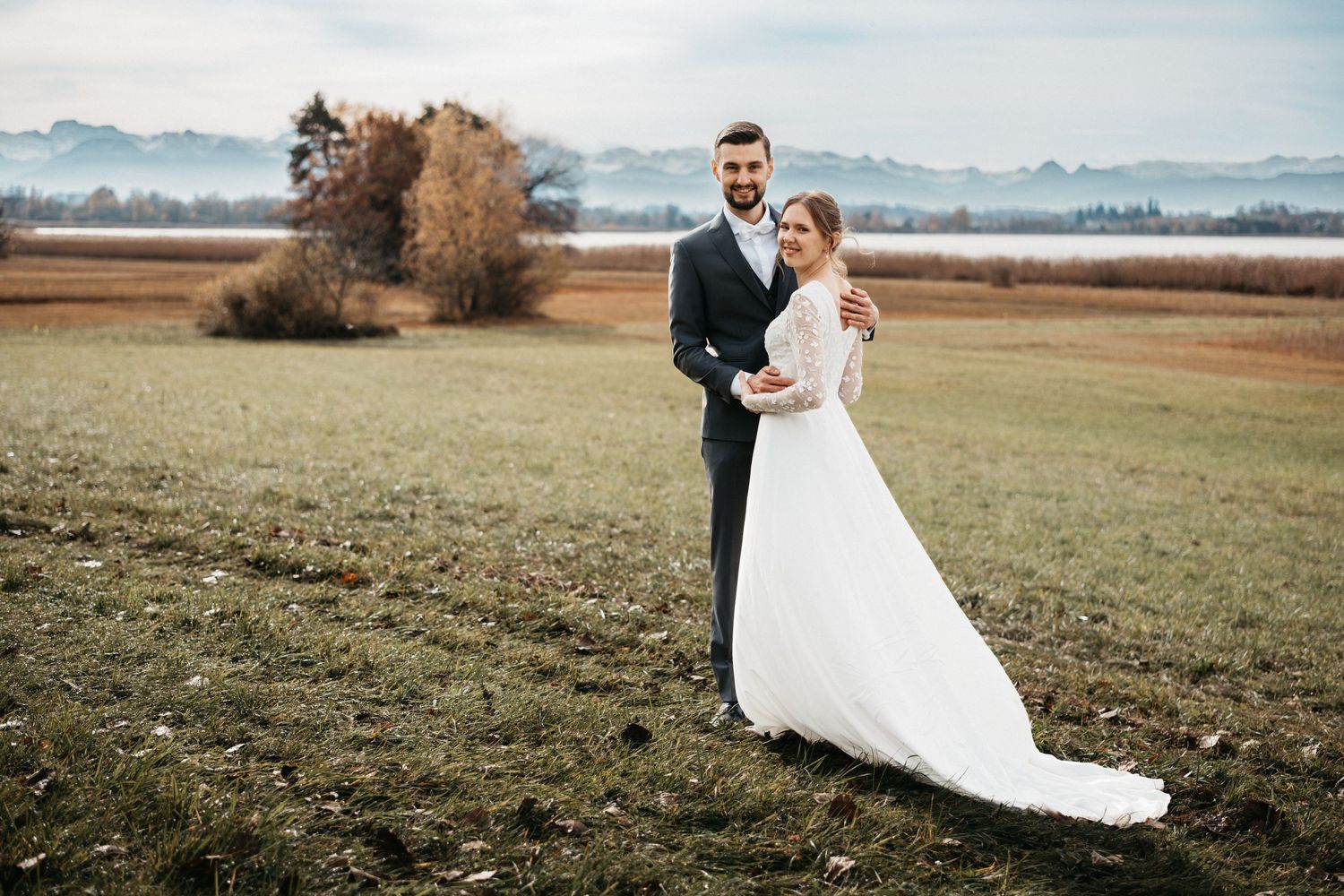 A couple in wedding attire stands together in a grassy field with mountains visible in the misty background.