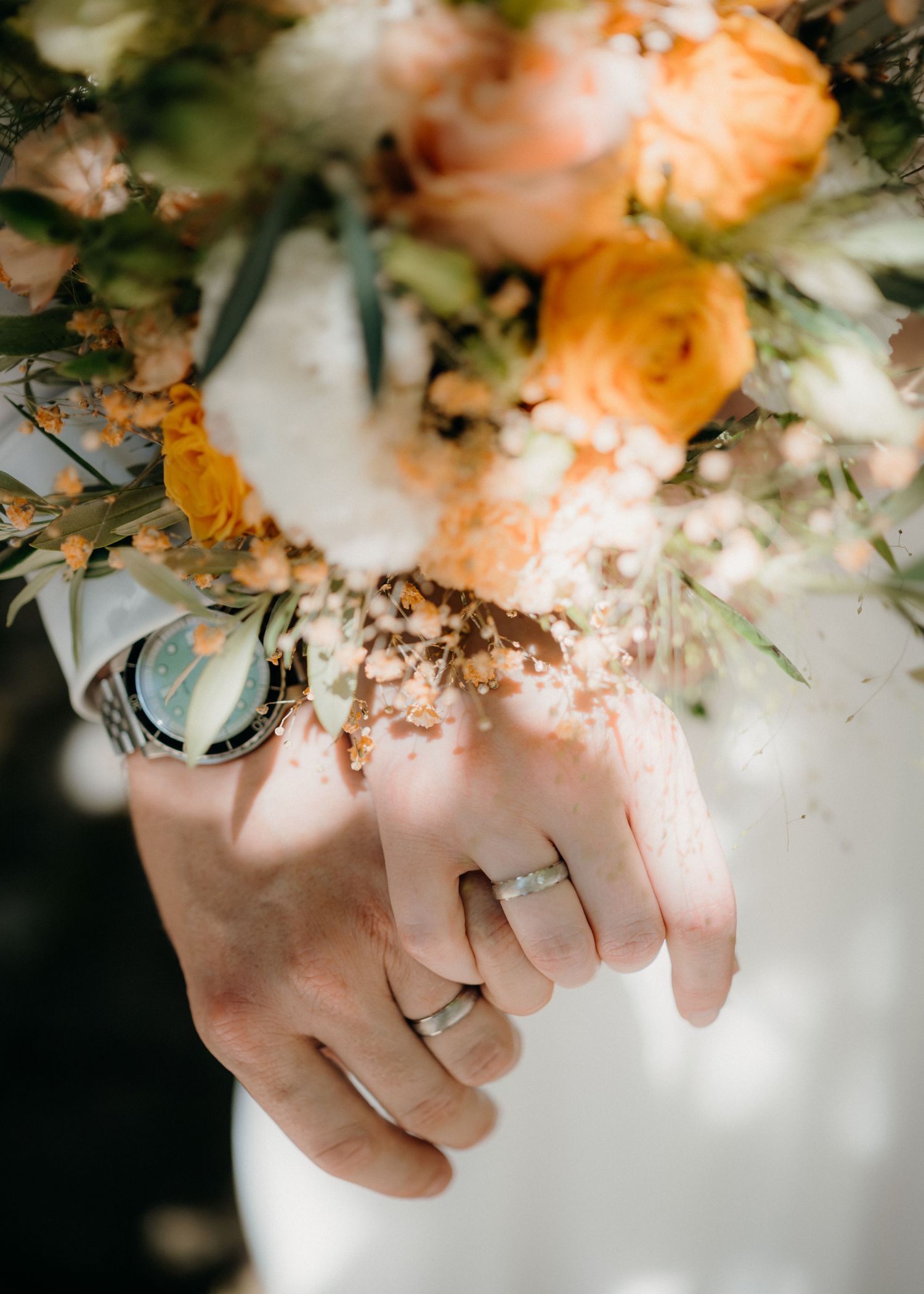 Newlywed couple holding hands with wedding rings, a watch, and an orange and white floral bouquet overhead.