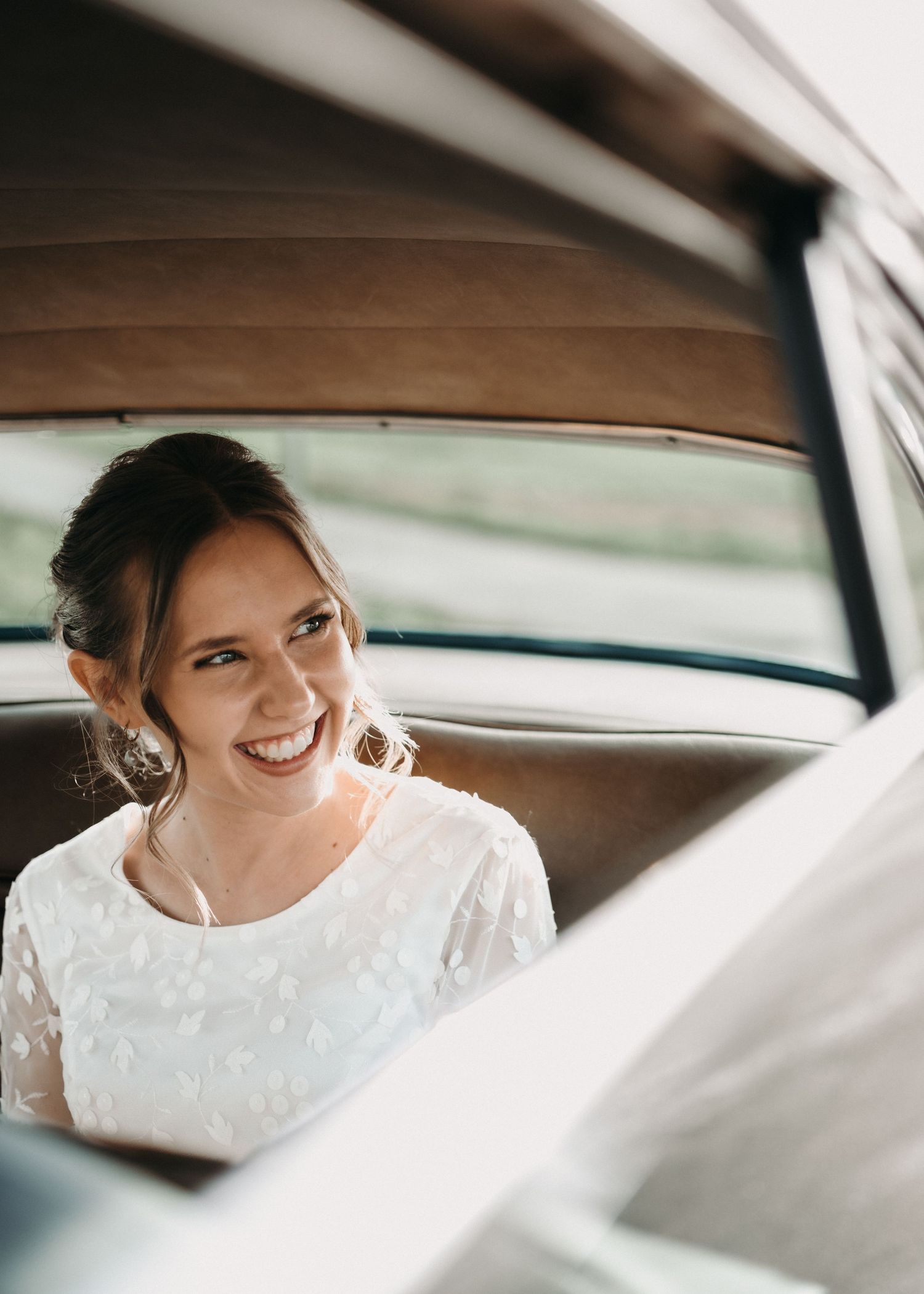 A bride in a white lace wedding dress smiles joyfully while sitting in a vintage car interior.