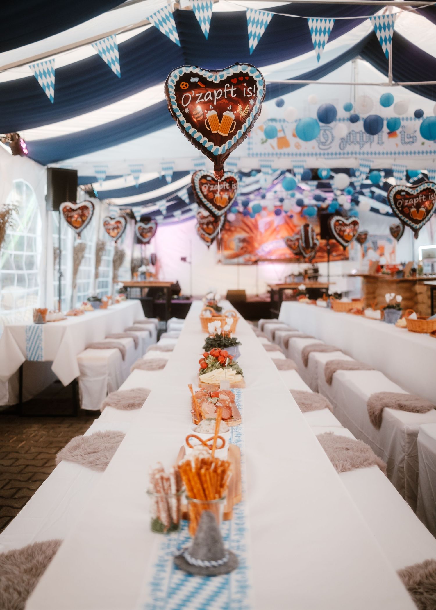 Long white tables decorated with Bavarian blue and white banners and heart-shaped balloons set up for an Oktoberfest celebration.