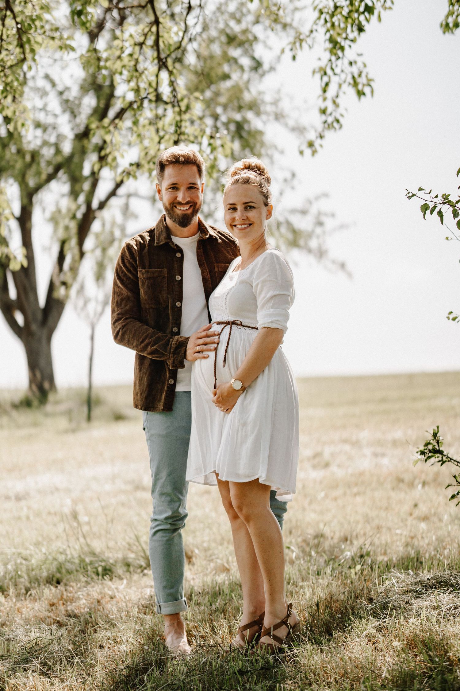 A couple shares a joyful moment together in a sun-dappled meadow beneath trees on a bright afternoon.