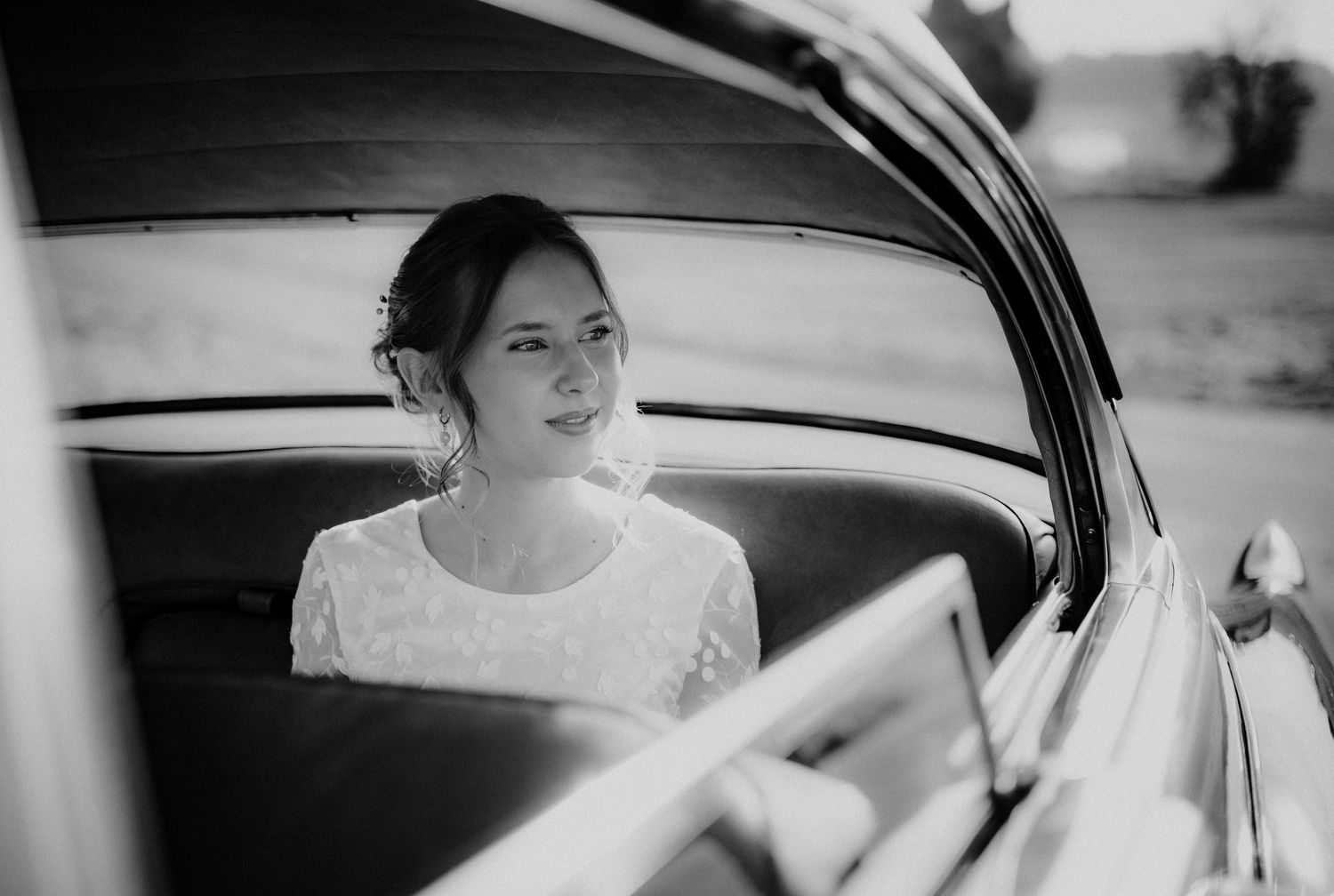 Black and white photo showing someone in a lace wedding dress sitting inside a vintage car.