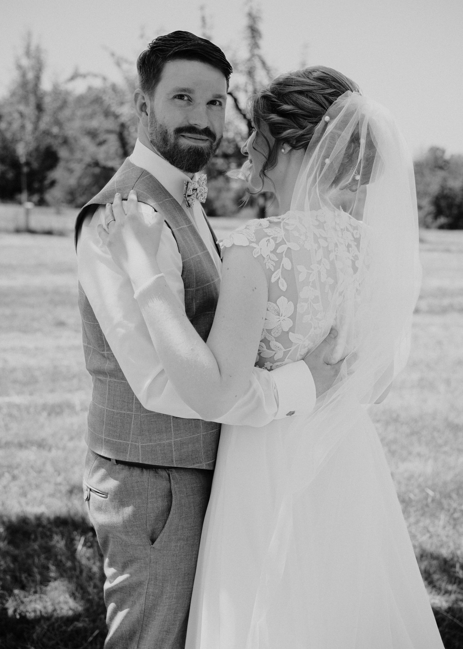 Black and white wedding photo of a bearded groom in a suit embracing a bride in a lace dress and veil outdoors.