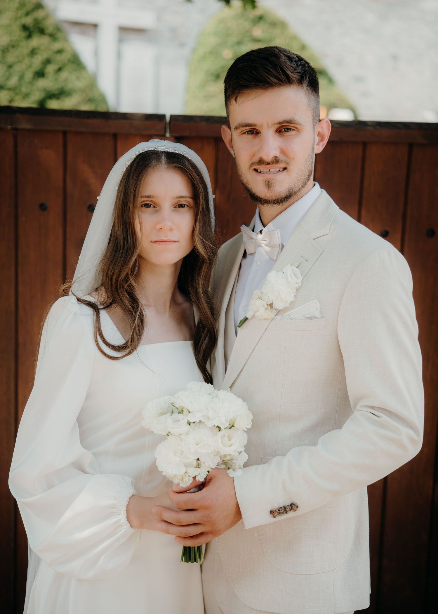 A bride and groom pose together on their wedding day, she in a white gown with veil, he in a cream suit.