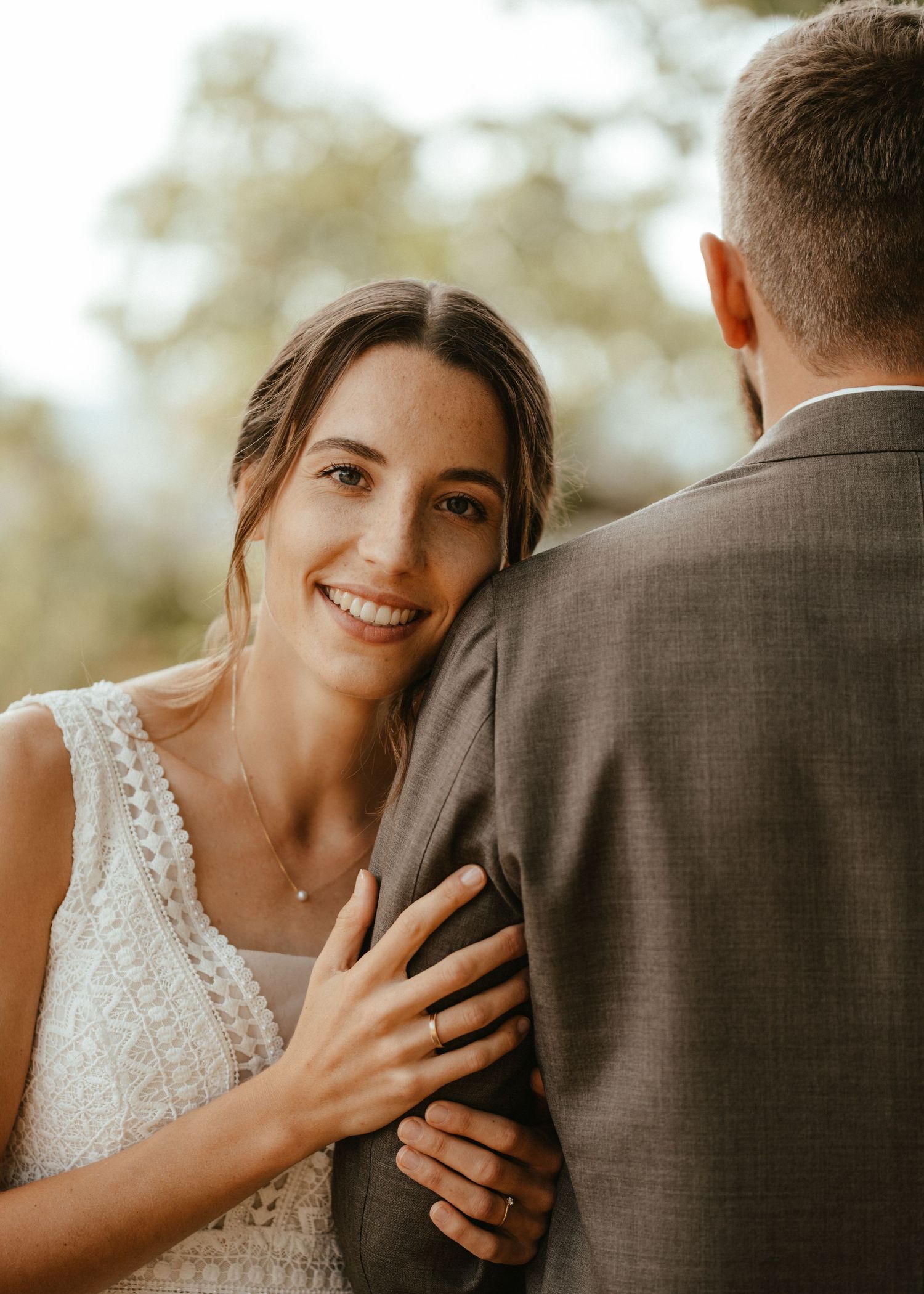 Smiling bride in white lace dress rests her hand on groom's gray suit jacket during outdoor wedding ceremony.