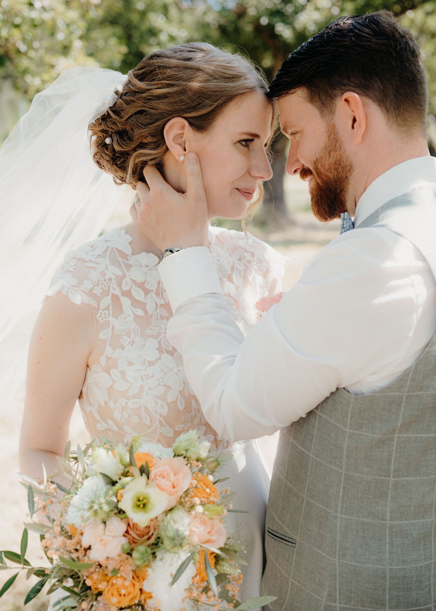 A bride and groom share an intimate moment, touching foreheads, as she holds a pastel bouquet in a lace wedding gown.