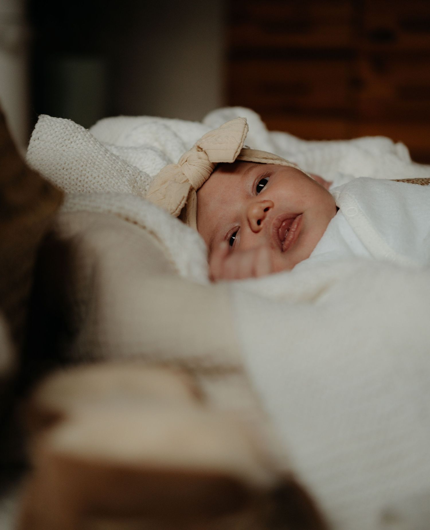 Baby wearing white knit sweater lies on soft bedding while looking towards the camera with a gentle expression.