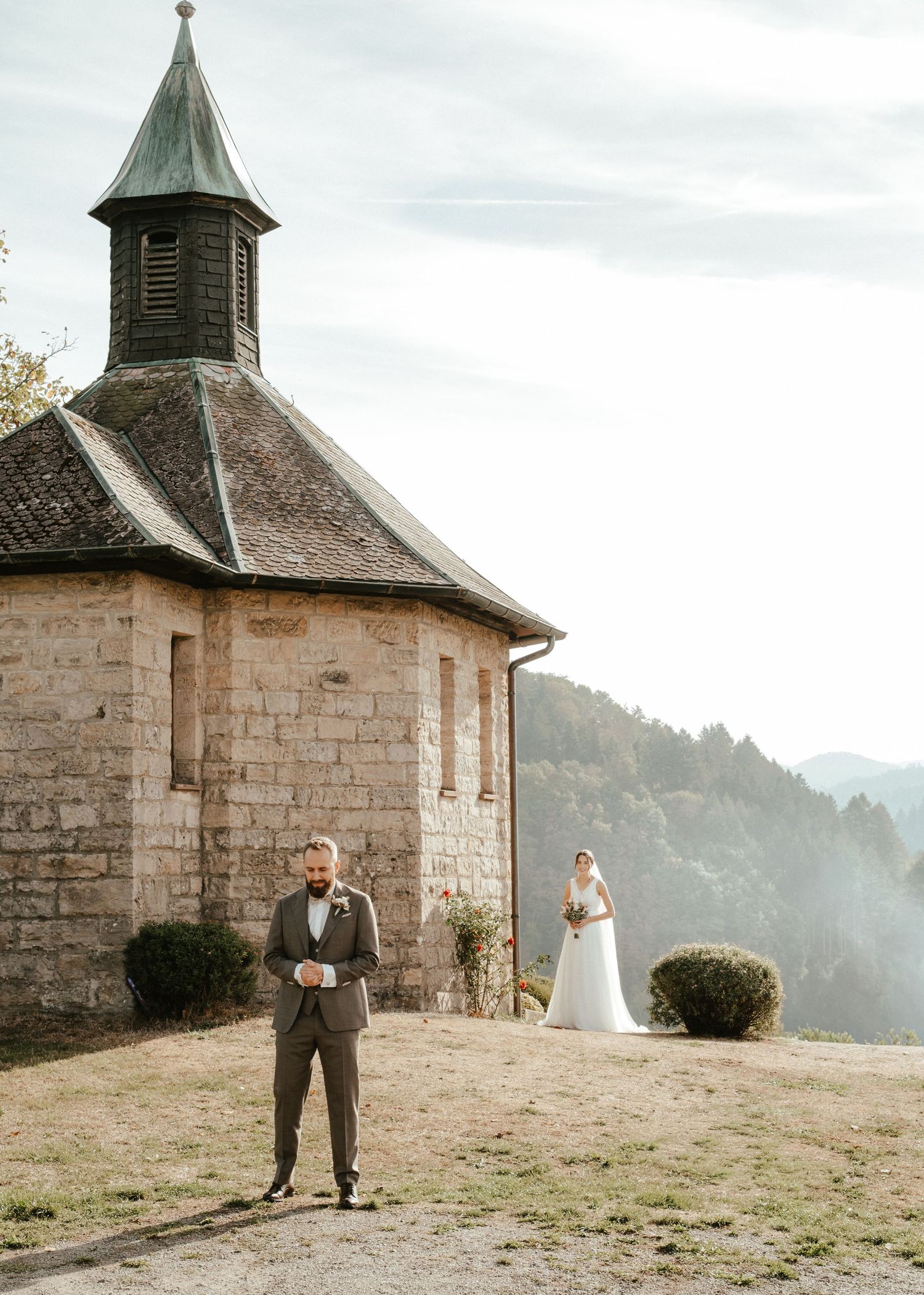 Bride and groom pose outside a historic stone chapel with mountain views on their wedding day.