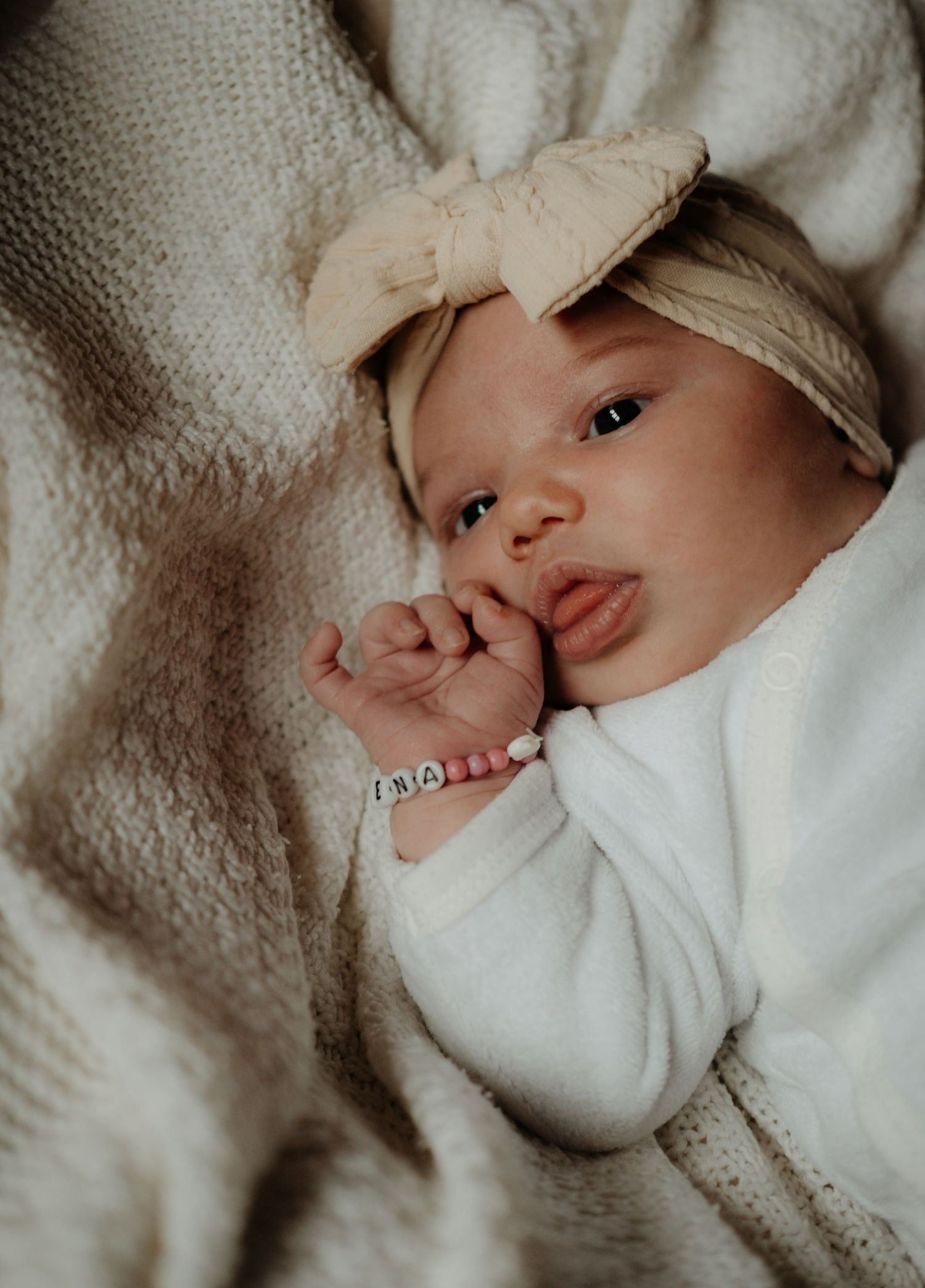 Baby wearing a cream colored bow headband lies on textured blanket while making an adorable expression.